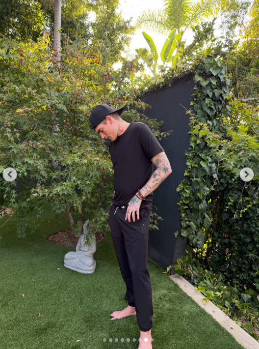 Presley Gerber looks down as he adjusts his black pants, standing barefoot in the garden with his backward cap and tattoos visible against the lush greenery. | Source: Instagram/presleygerber