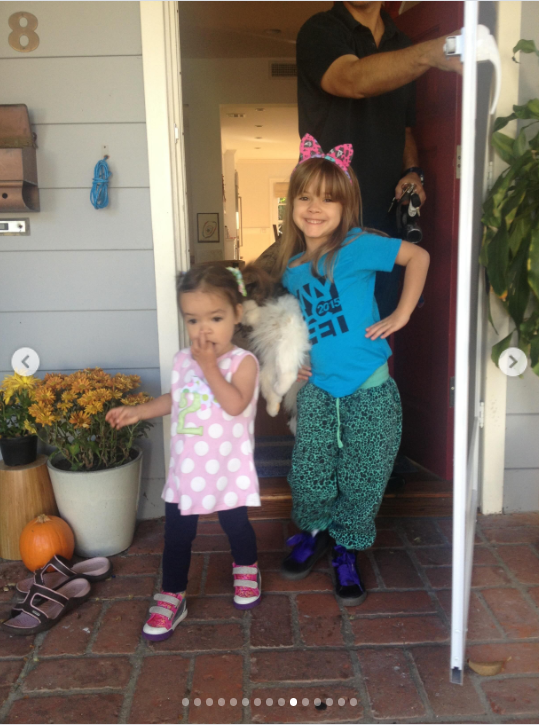 Then comes a childhood holiday memory. The girls stand outside a home, apparently dressed for Halloween, with Mabel wearing a bright blue T-shirt, patterned green pants, and a large pink bow headband. Her younger sister wears a pink-and-white polka-dot dress, dark leggings, and pink sandals. Pumpkins and other seasonal details are visible around the doorway. | Source: Instagram/mabellowe_