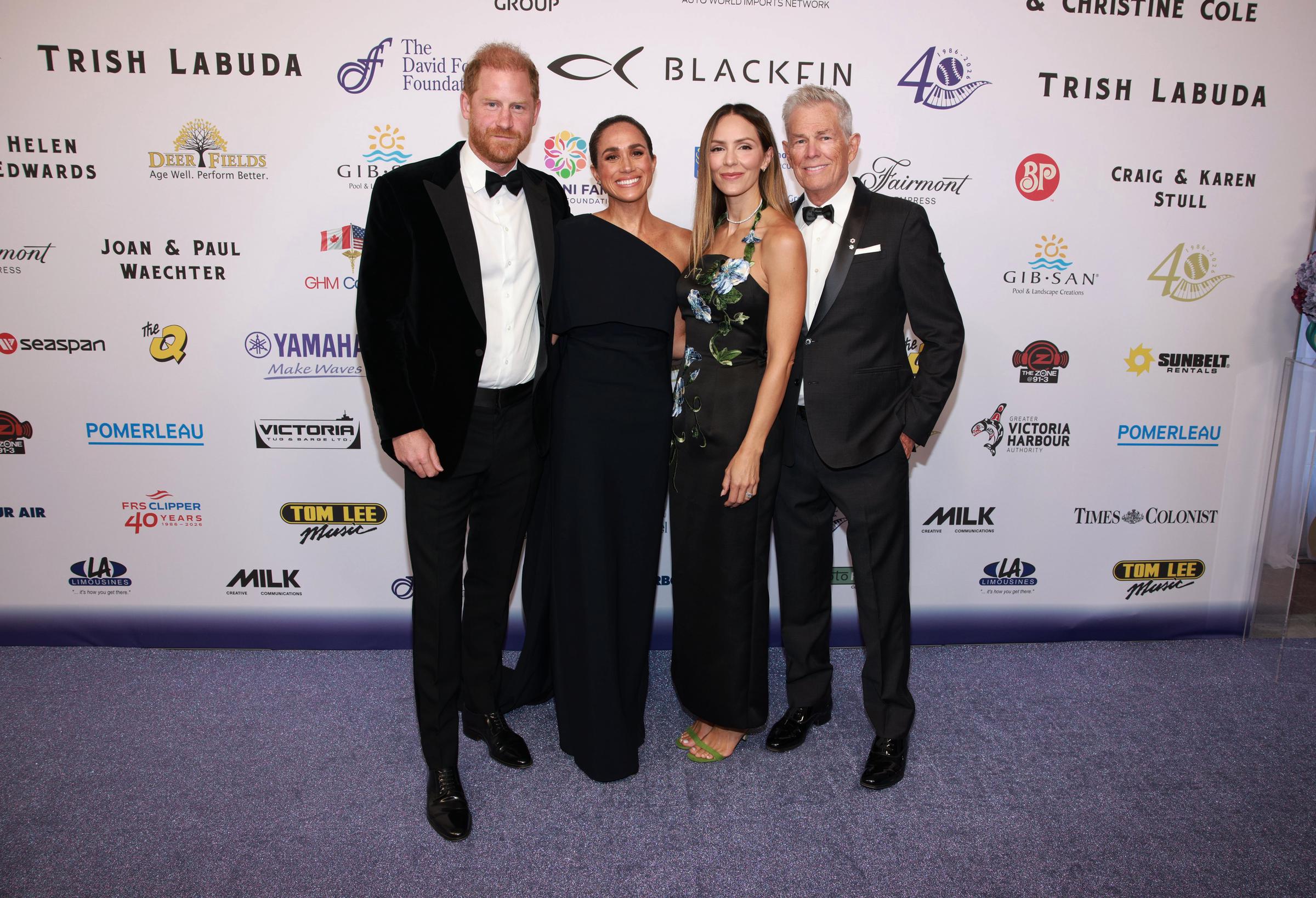 Prince Harry and Meghan Markle pose with fellow guests at a formal event, wearing black-tie attire in front of a sponsor-covered backdrop. | Source: Getty Images