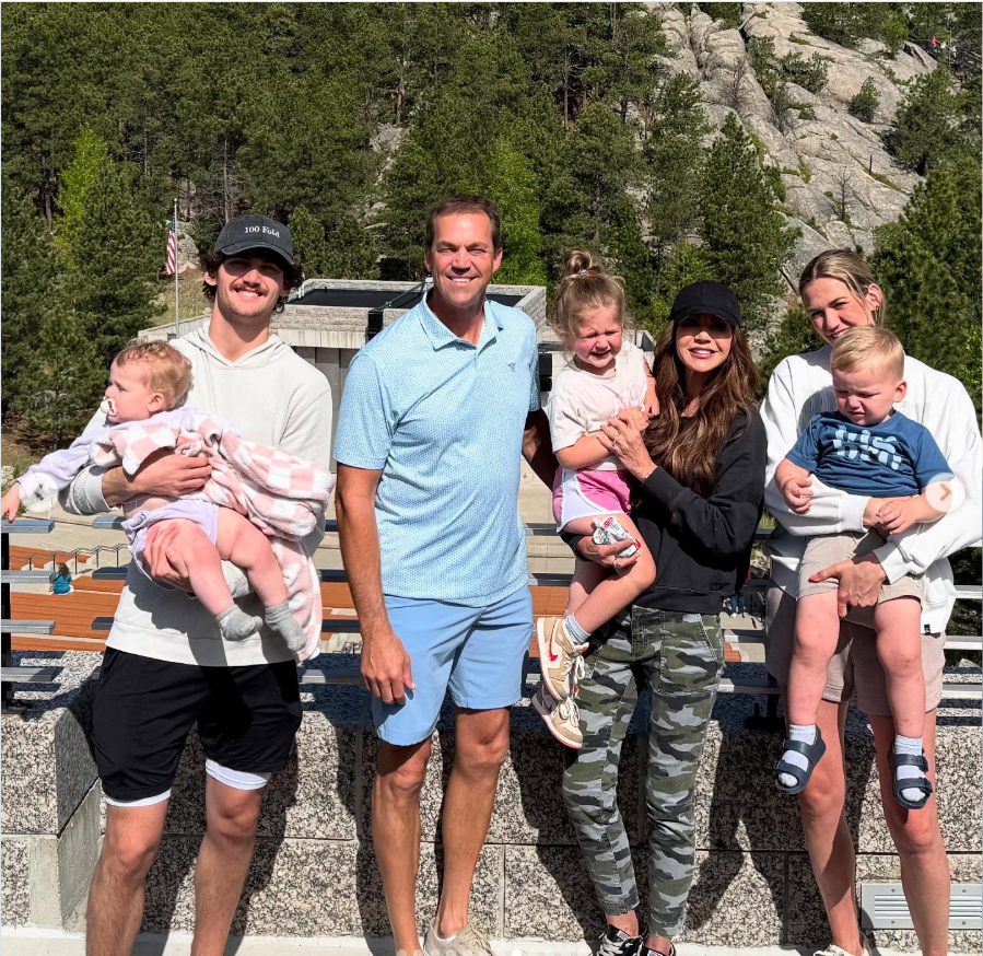 Bryon and Kristi Noem pose with family members outdoors, smiling as they stand together with children against a scenic backdrop of trees and rocky hills. | Source: Instagram/kristinoem