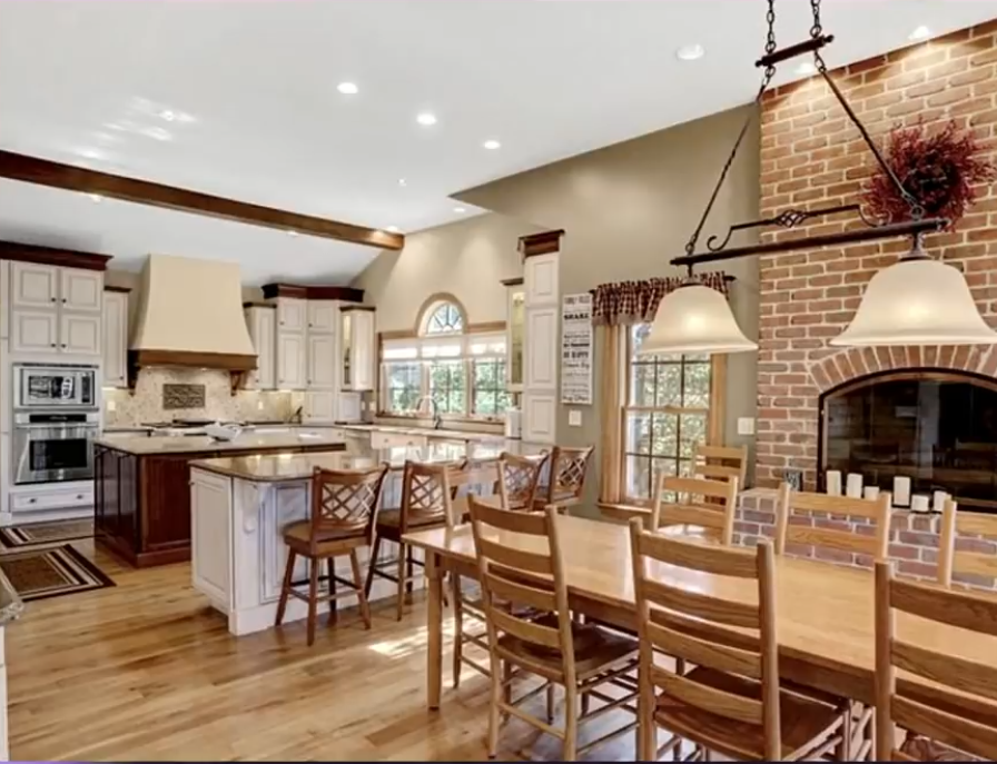 The open kitchen and dining area features cream cabinets, wooden floors, exposed ceiling beams, and a prominent brick fireplace. A long wooden dining table sits beneath two pendant lights, with matching chairs arranged around it.