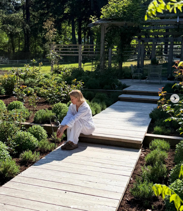 Pamela Anderson rests on the steps of a bleached wooden boardwalk at Arcady, sitting barefoot in loose white linens amid manicured boxwood shrubs and blooming flower beds. Looking toward the greenery before a sheltered wooden pergola, she captures the quiet satisfaction of self-reliance, captioning the post, "I think of life as a garden… You get to replant, start again, and make it yours. 🤍 Love, P". | Source: Instagram/pamelaanderson