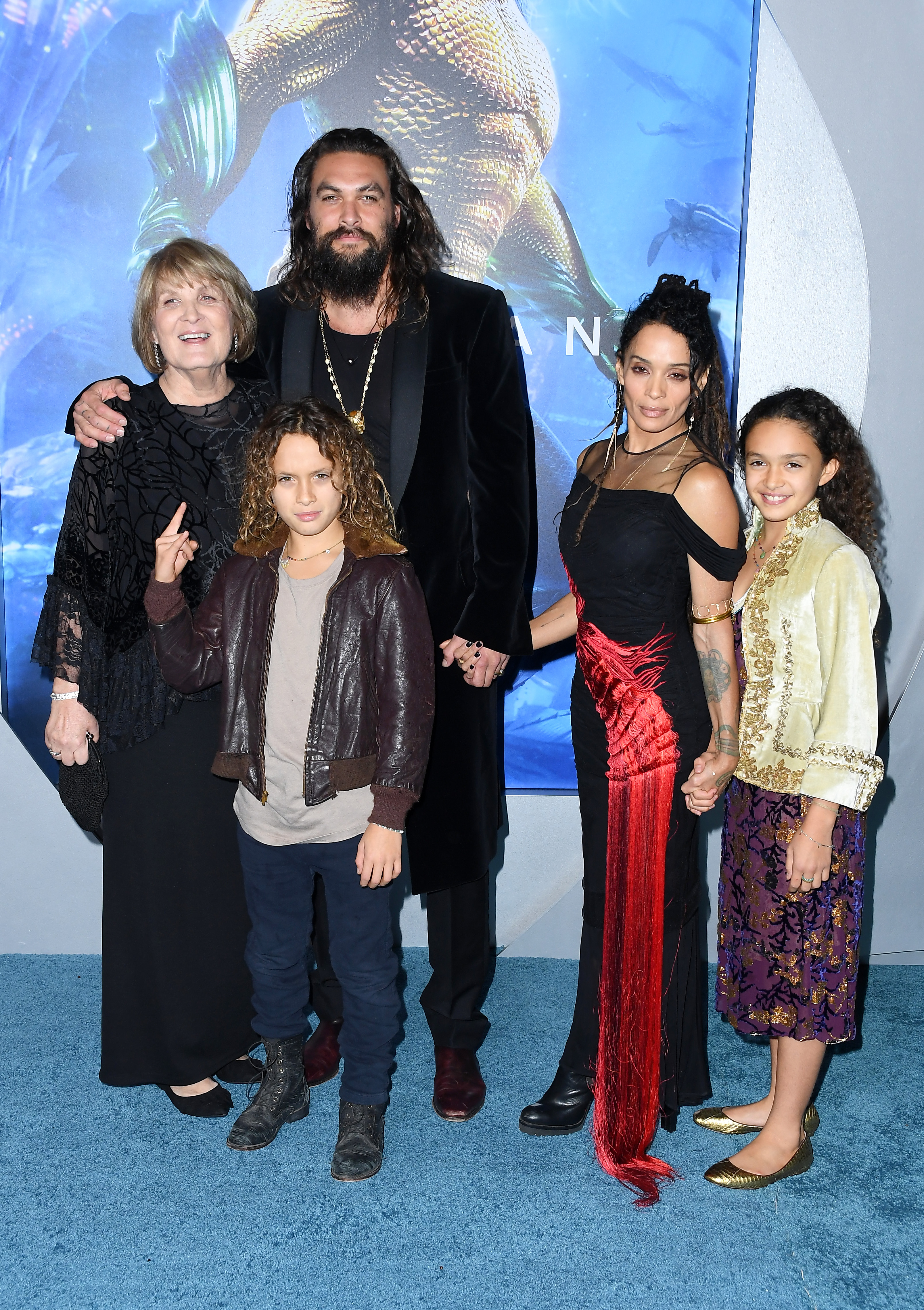Jason Momoa is joined by his mother Coni Momoa, son Nakoa-Wolf Manakauapo Namakaeha Momoa, partner Lisa Bonet, and daughter Lola Iolani Momoa on the blue carpet at the Los Angeles premiere of Warner Bros. Pictures' "Aquaman" at the TCL Chinese Theatre on December 12, 2018, in Hollywood, California. Momoa wears a black velvet tuxedo coat with gold statement necklaces while posing proudly with his family, including Bonet in a black evening gown accented with a cascading vibrant crimson fringe sash. | Source: Getty Images