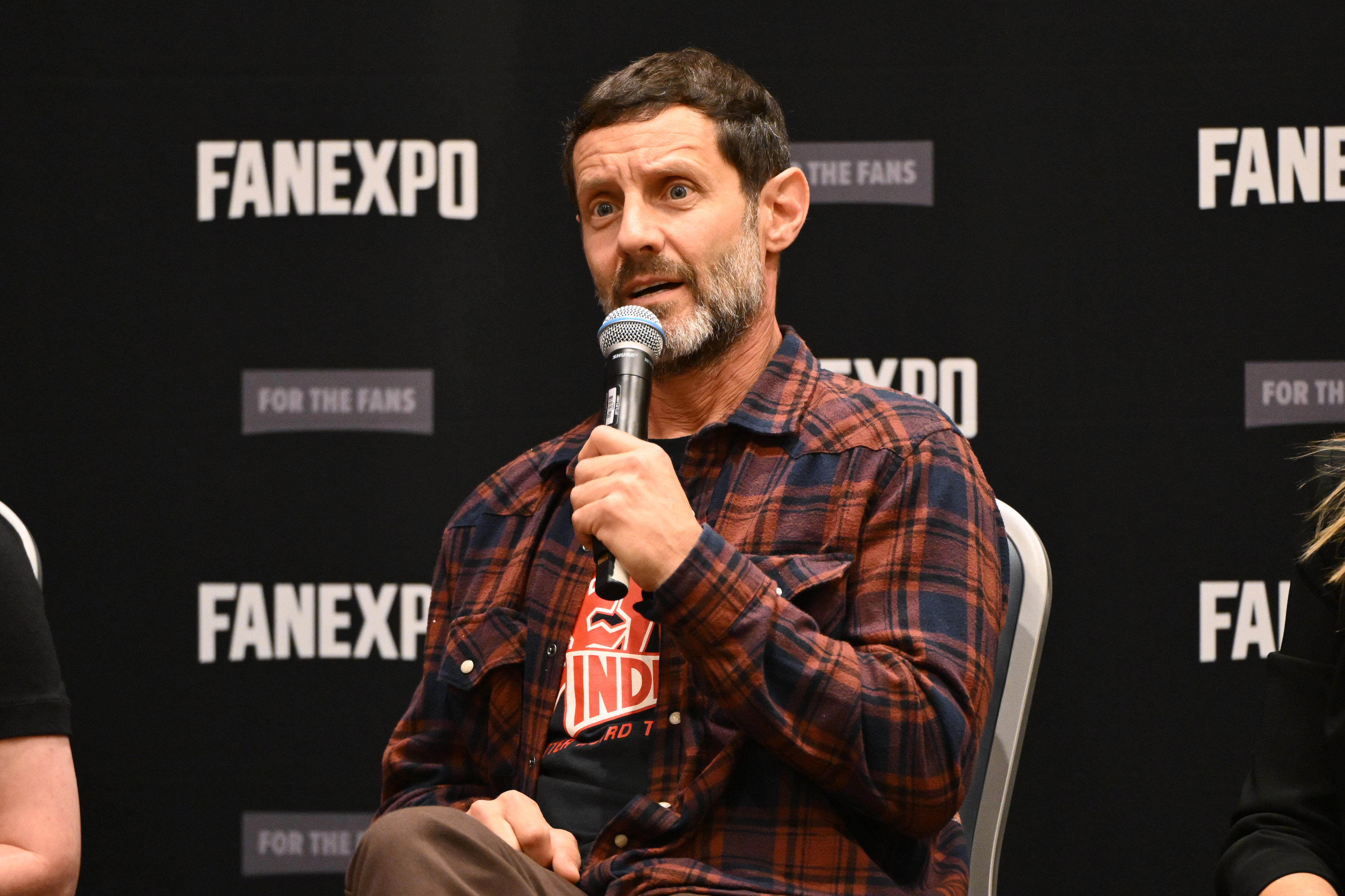 Omri Katz presents a distinguished, mature look with prominent silver throughout his full beard, short dark hair, and expressive, weathered features that emphasize his transformation far beyond his boyish teen years, wearing a rustic plaid flannel over a graphic tee. The photo captures him speaking on stage into a microphone during FanExpo Chicago at the Donald E. Stephens Convention Center in Rosemont, Illinois, on August 15, 2026.