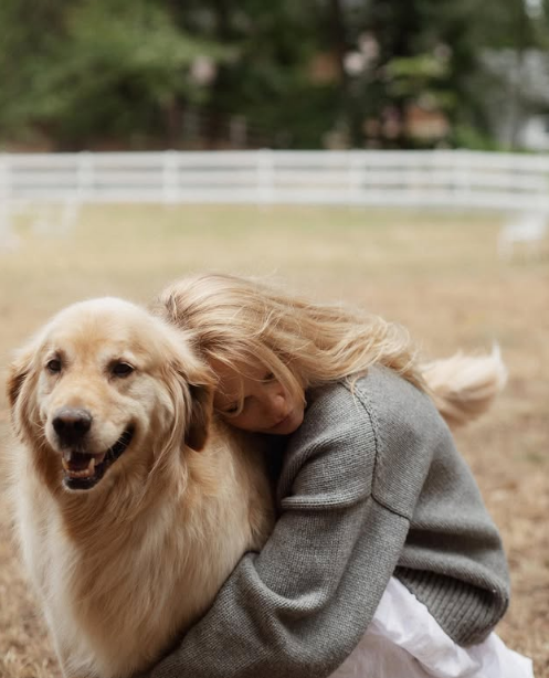 Pamela Anderson buries her face into the soft fur of her golden retriever during a quiet moment outdoors on her Arcady grounds. Dressed warmly in a chunky grey knit sweater and a flowing white skirt against the backdrop of trees and white paddock fencing, she shares a tender embrace that highlights her deep bond with animals and the comforting simplicity of home on Vancouver Island. She posted the heartwarming photo alongside the caption, "True love never ends… 🤍". | Source: Instagram/pamelaanderson