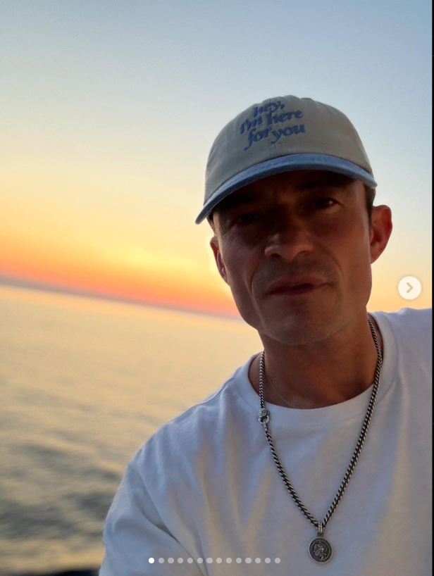 Orlando Bloom in a white crew neck shirt, with a thin chain around his neck and a baseball cap reading "hey, i'm here for you." From an Instagram post dated July 9, 2025. | Source: Instagram/orlandobloom