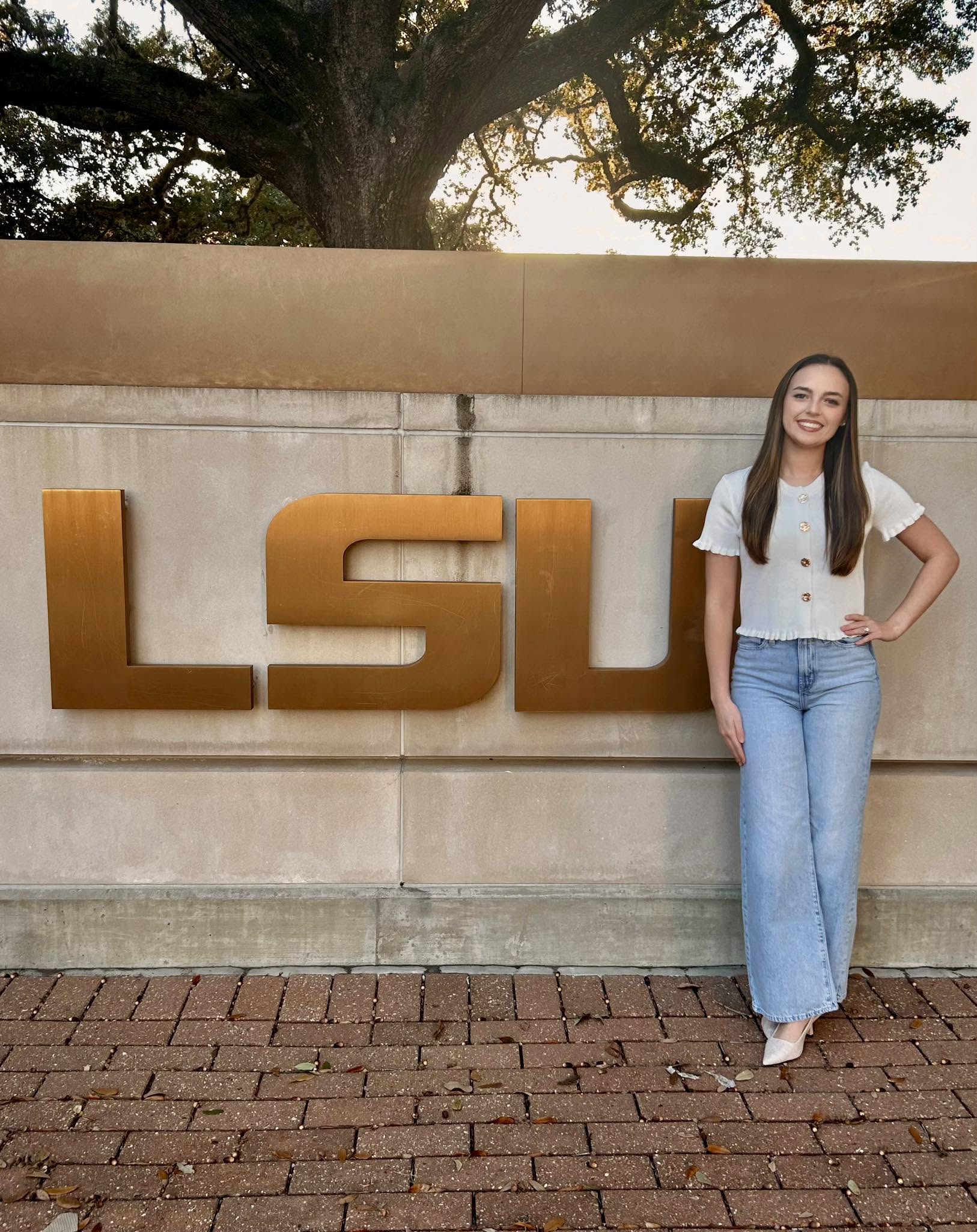 Leah Victoria Fonte poses proudly in front of a large metallic LSU sign on the university campus. The senior biological sciences major smiles with one hand on her hip, dressed casually yet stylishly in a white short-sleeved button-front top, wide-leg light-wash denim jeans, and pointed heels. | Source: Facebook/Leah Fonte