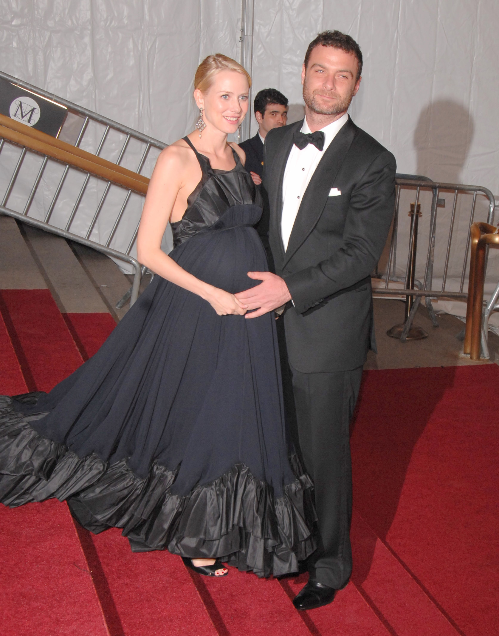 Naomi Watts and Liev Schreiber pose together on the red carpet at a formal event. Watts wears a flowing black gown that highlights her baby bump. Schreiber stands beside her in a classic tuxedo, resting a hand gently on her belly.