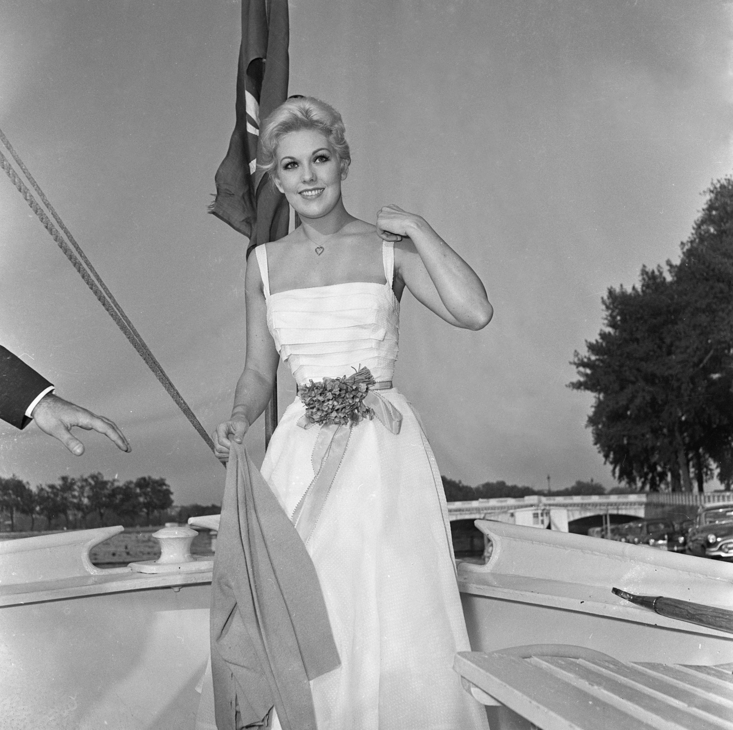 Kim Novak poses in a white sleeveless gown while aboard the Borde-Brétigny on the Seine in Paris, smiling beside a flag. | Source: Getty Images