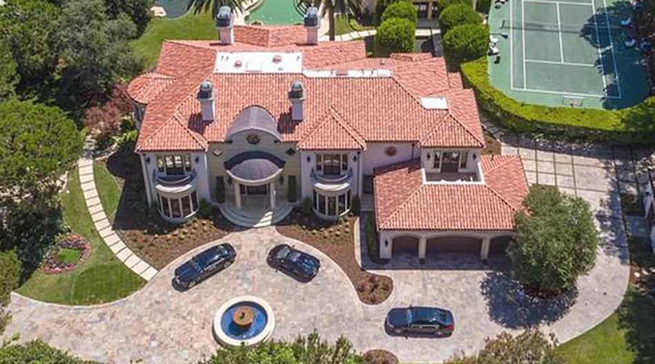 An elevated view displays the estate's sprawling Italianate architecture, terracotta tile roofs, and attached garage wing, alongside the wide cobblestone motor court fountain and neighboring tennis court. | Source: YouTube/Taste of Country