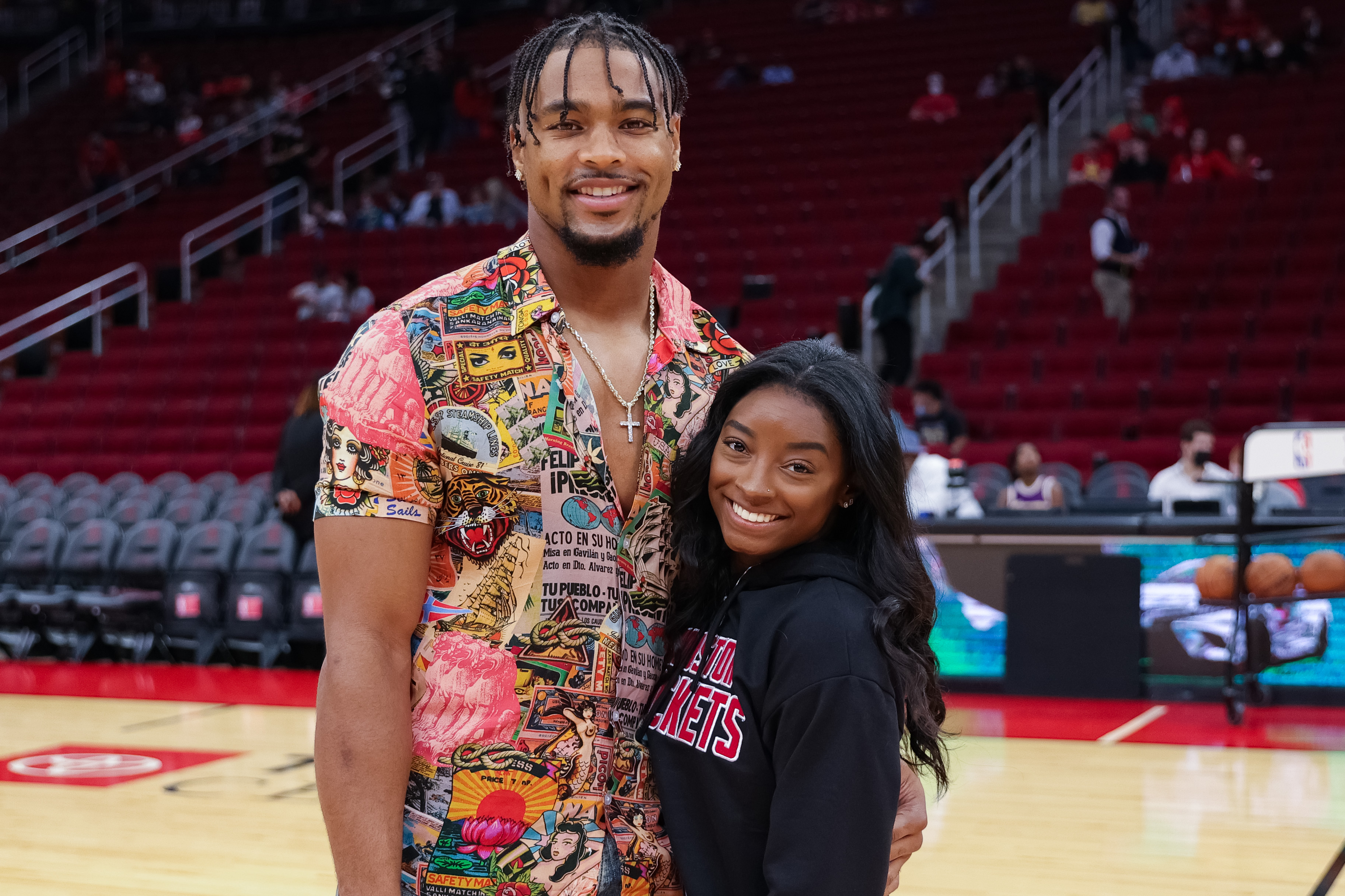 Simone Biles and Jonathan Owens pose courtside during a game between the Houston Rockets and the Los Angeles Lakers at the Toyota Center in Houston, Texas, on December 28, 2021. Biles smiles in a black Houston Rockets hoodie, while Owens stands beside her in a colorful printed shirt with layered chains.