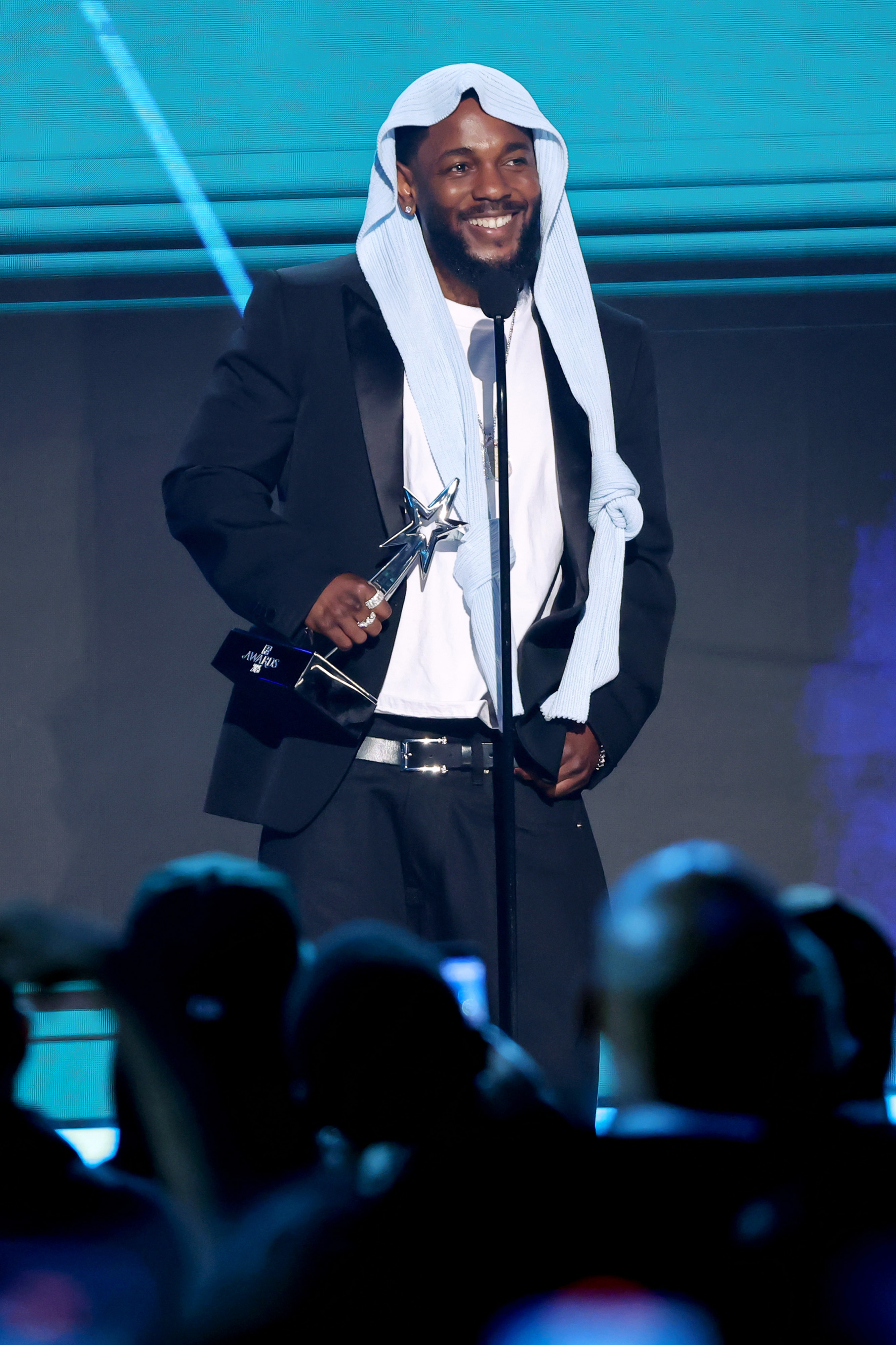 Kendrick Lamar flashes a bright smile while holding a trophy onstage at the 2025 BET Awards. Dressed in his now-iconic head covering and black suit, the rapper appeared genuinely moved by the evening's continued recognition.
