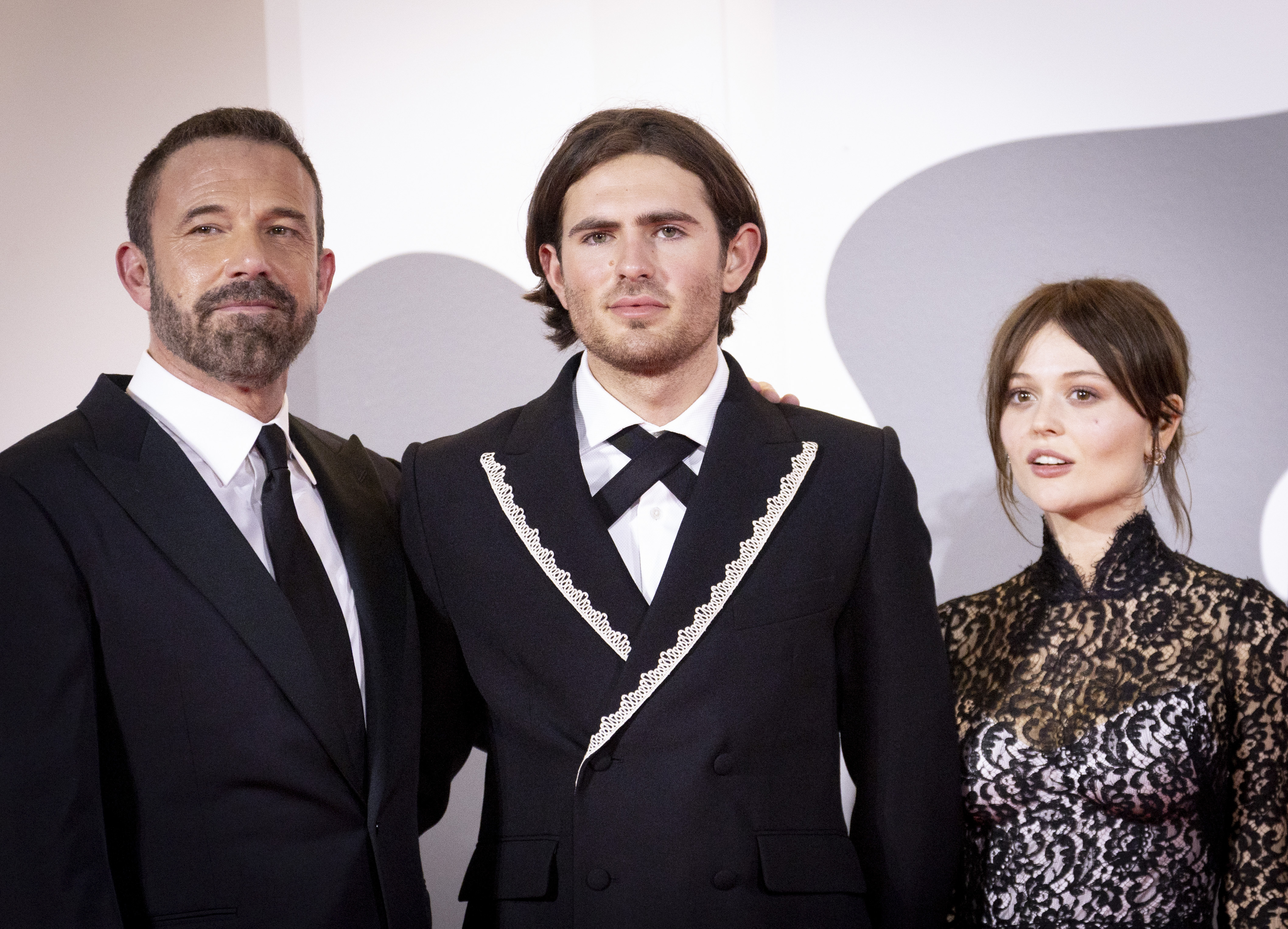 Commanding the center of the frame, Indiana Affleck brought an artistic flair to the red carpet in an asymmetrical black wrap jacket trimmed with ornate white braided embroidery along the lapels. A crossed black cravat-style necktie over a stiff white collar, paired with parted shoulder-length hair and subtle stubble, lent an eclectic, modern energy to his festival appearance alongside uncle Ben Affleck and co-star Emily Alyn Lind at the "Company" screening during the 83rd Venice International Film Festival on September 8, 2026. | Source: Getty Images