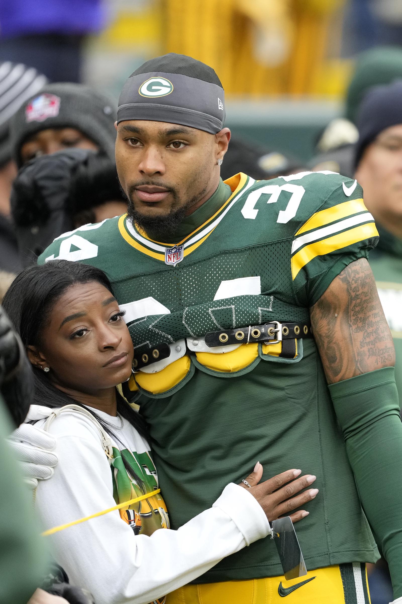 Simone Biles embraces Jonathan Owens on the sidelines before kickoff at Lambeau Field on October 29, 2023. Biles rests her head against Owens' chest as he stands in uniform, both appearing serious and focused ahead of the Green Bay Packers' game against the Minnesota Vikings.