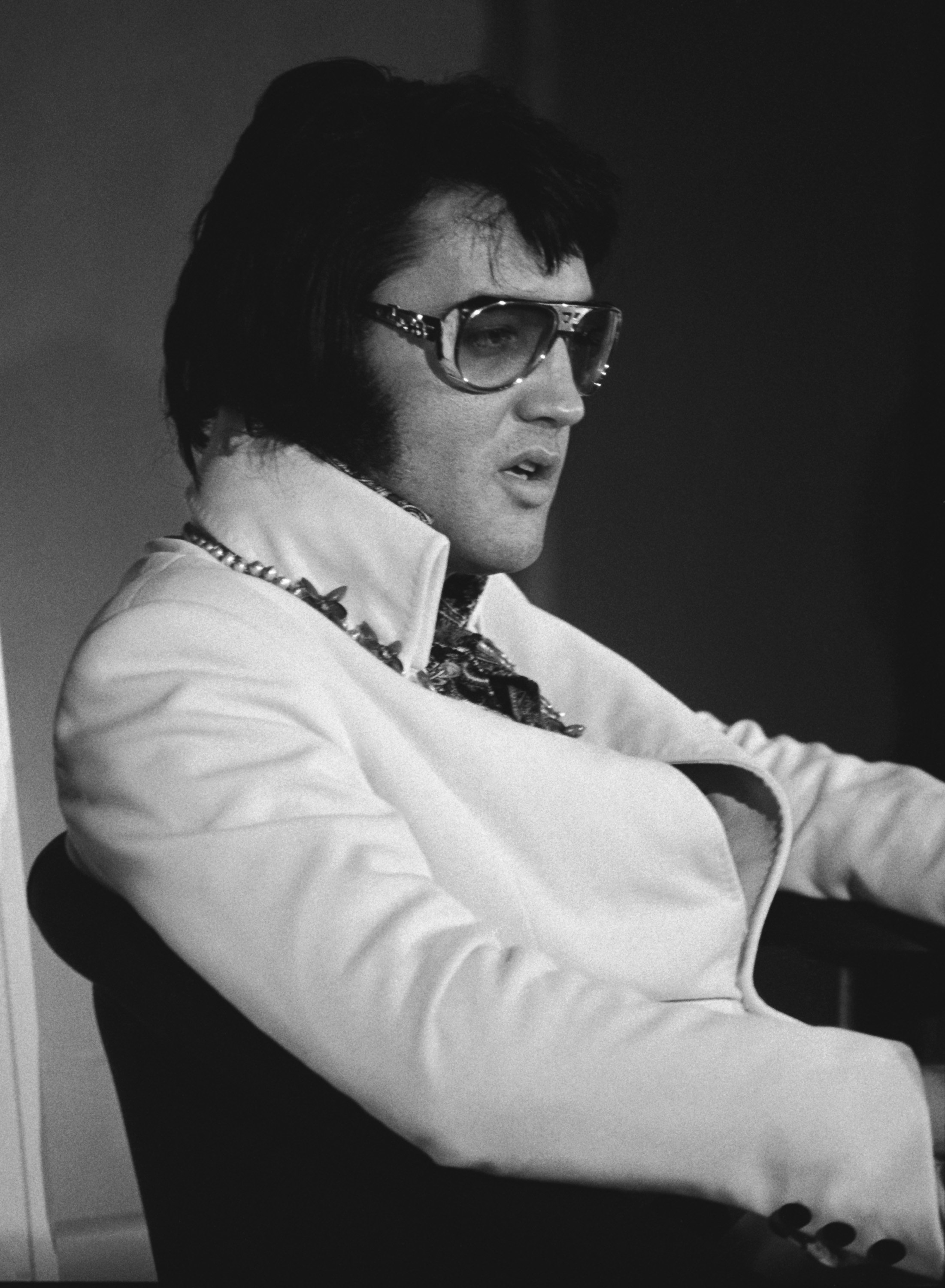 Elvis Presley participates in a promotional interview at the Las Vegas Hilton in Las Vegas, Nevada, on September 4, 1972, for his upcoming televised concert "Elvis: Aloha from Hawaii", wearing an open, high-collared light jacket over an ornate patterned ascot and chain necklace, paired with his signature oversized tinted aviator sunglasses and sideburns