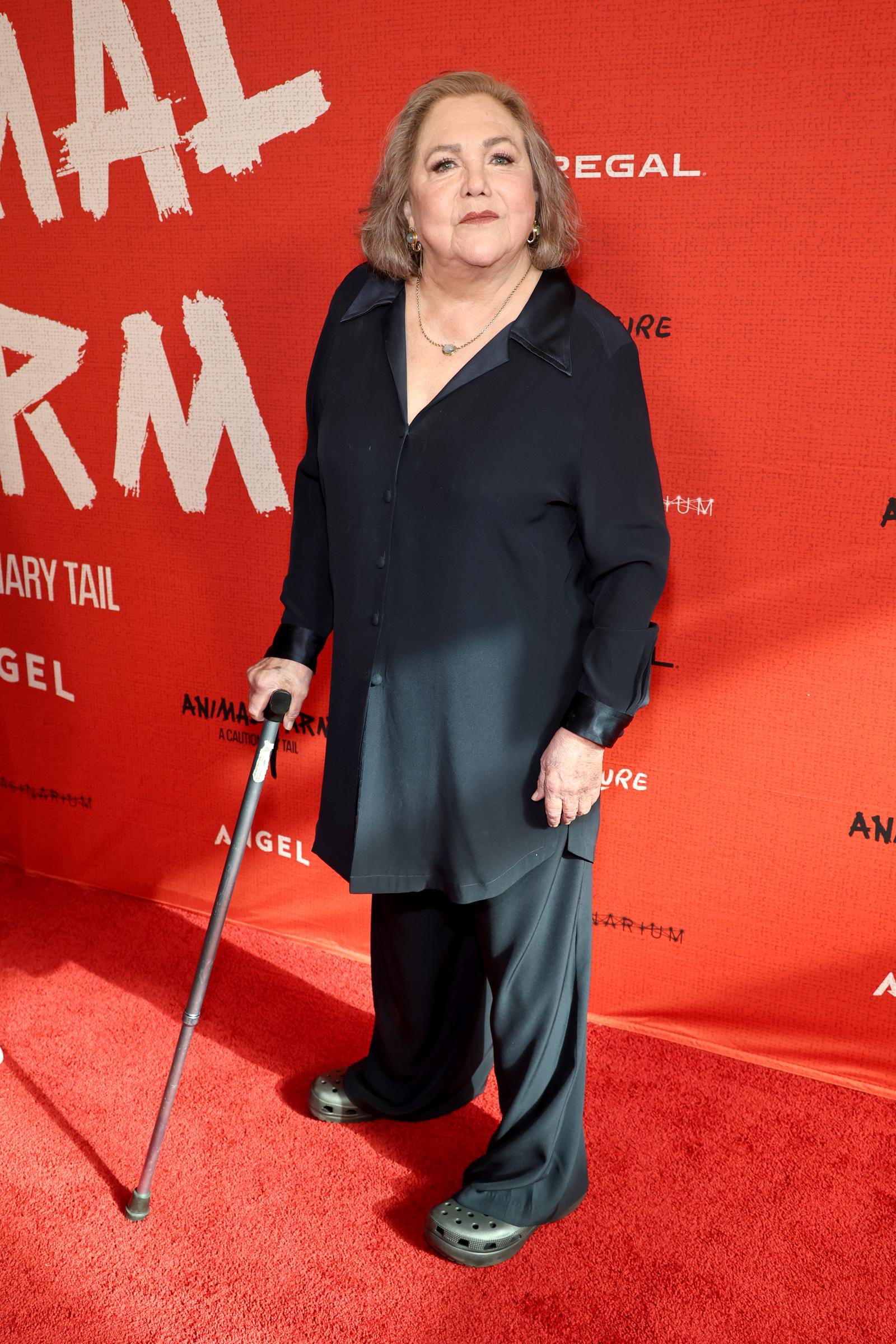 Kathleen Turner arrives at the "Animal Farm" premiere in a loose-fitting navy button-down top and matching trousers, accessorized with gold jewelry. The actress uses a cane as she poses on the red carpet.