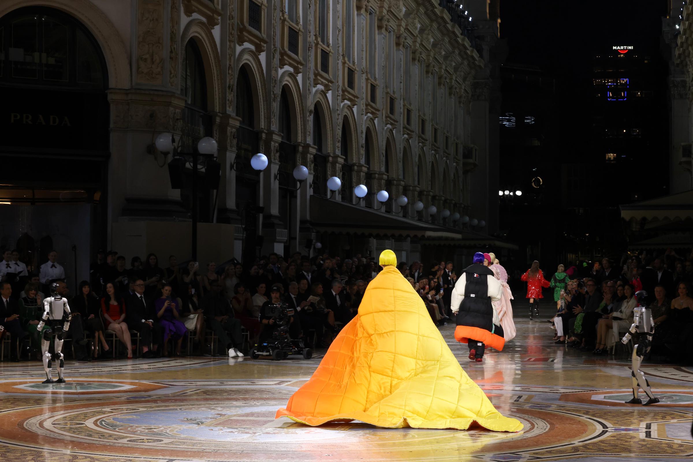 Bold, sculptural silhouettes met futuristic spectacle as models commanded the runway on September 22, 2026, in Milan, Italy. A sweeping canary-yellow and orange gradient quilted cape glided across the historic mosaic floor of Galleria Vittorio Emanuele II, sharing the path with walking humanoid robots as onlookers watched from the sidelines. | Source: Getty Images