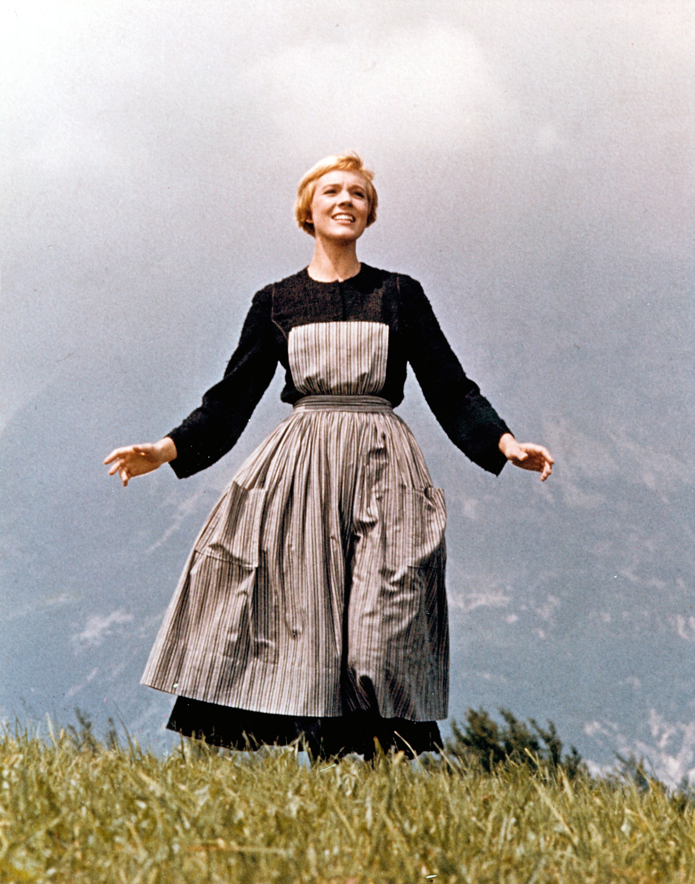 Julie Andrews delivers one of cinema's most celebrated opening sequences as Maria von Trapp in Robert Wise's 1965 masterpiece "The Sound of Music," filmed on location in the Austrian Alps in July 1964. Spinning joyfully across a grassy hilltop meadow against a hazy mountain backdrop, the actress sings with outstretched arms, dressed in her character's modest long-sleeved black dress layered with a striped apron pinafore.