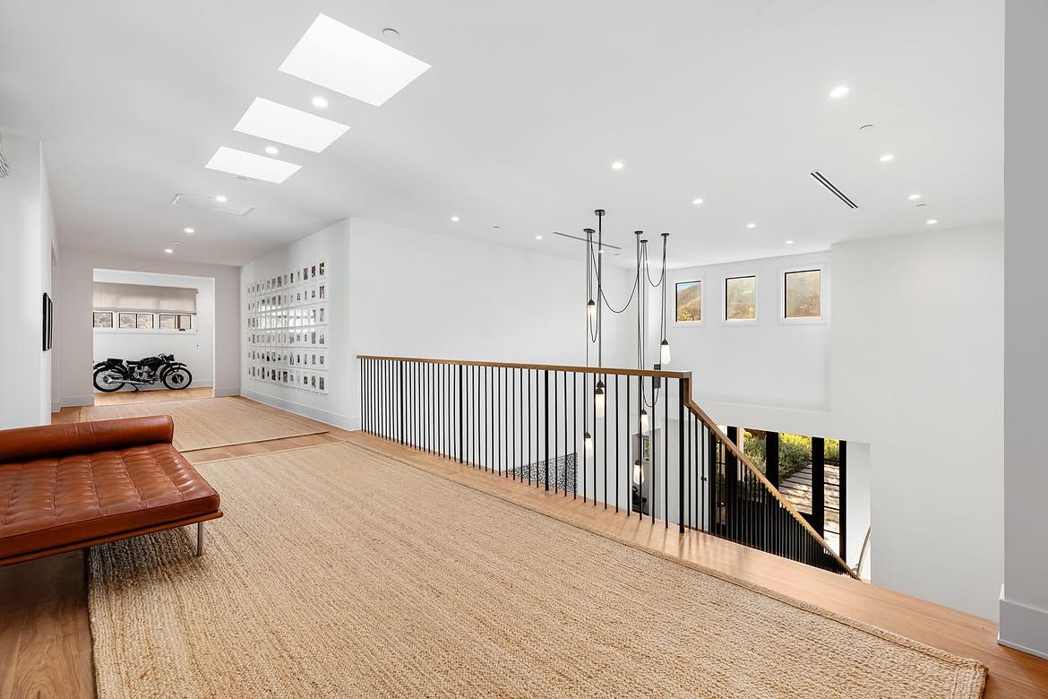 Skylights brighten the second-floor landing, where black railings and a wood handrail overlook the entry below. Down the hall, the motorcycle display appears again, showing how the unusual feature remains visible from several parts of the house. | Source: Instagram/tomer.fridman