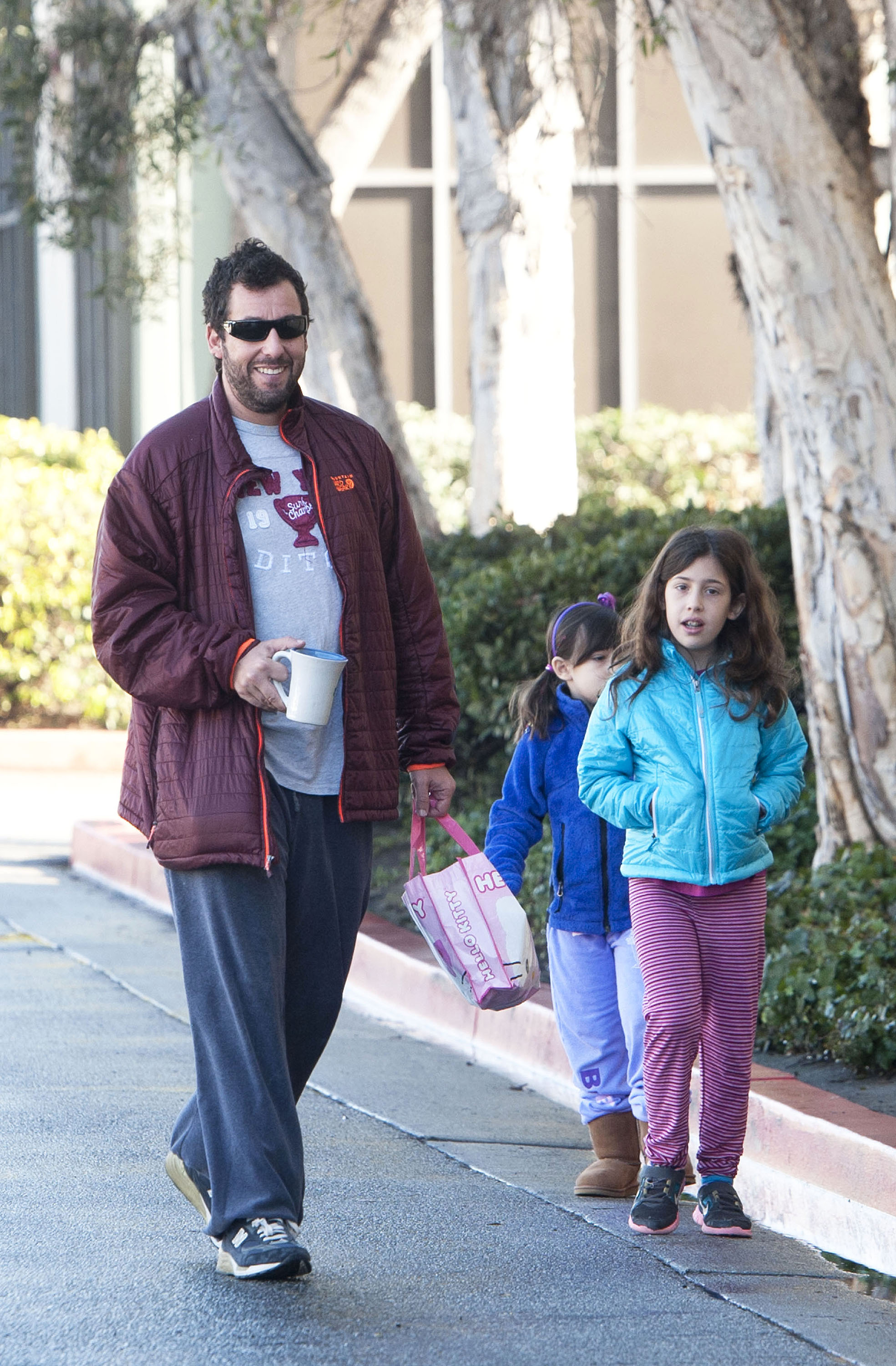 Adam Sandler steps out with daughters Sadie and Sunny in Los Angeles, casually walking them to dance class with a coffee in hand. Dressed in relaxed layers, he carries a small pink bag while the girls trail beside him in colorful jackets — capturing a laid-back, everyday family moment.