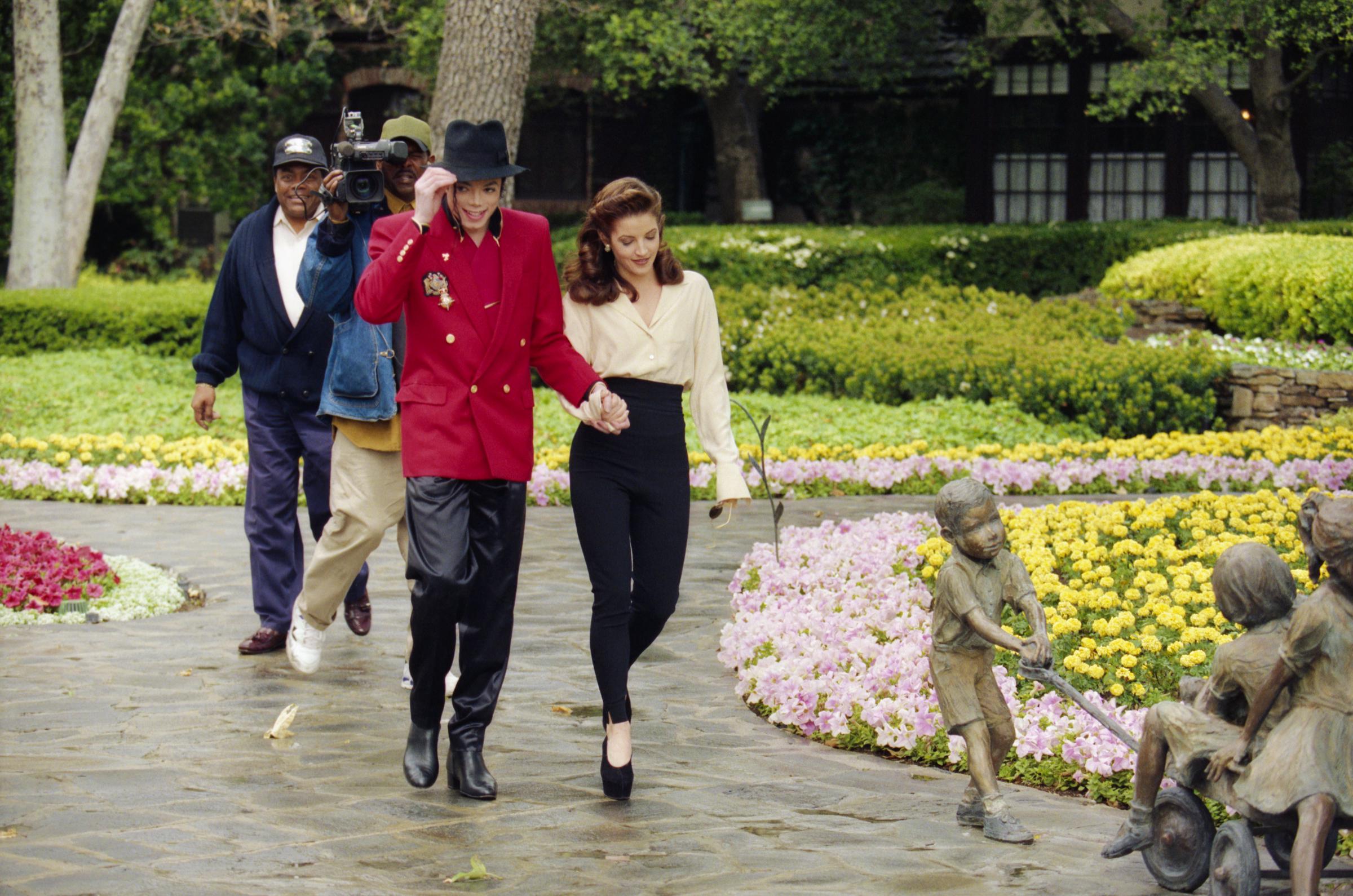 Lisa Marie Presley holds hands with her husband Michael Jackson as they stroll through the floral grounds of Neverland Ranch in preparation for the Children's World Summit on April 18, 1995, wearing an ivory long-sleeved silk blouse tucked into high-waisted black tapered trousers paired with heels, while Jackson adjusts his black fedora, dressed in a vibrant red double-breasted military-style blazer and black trousers.
