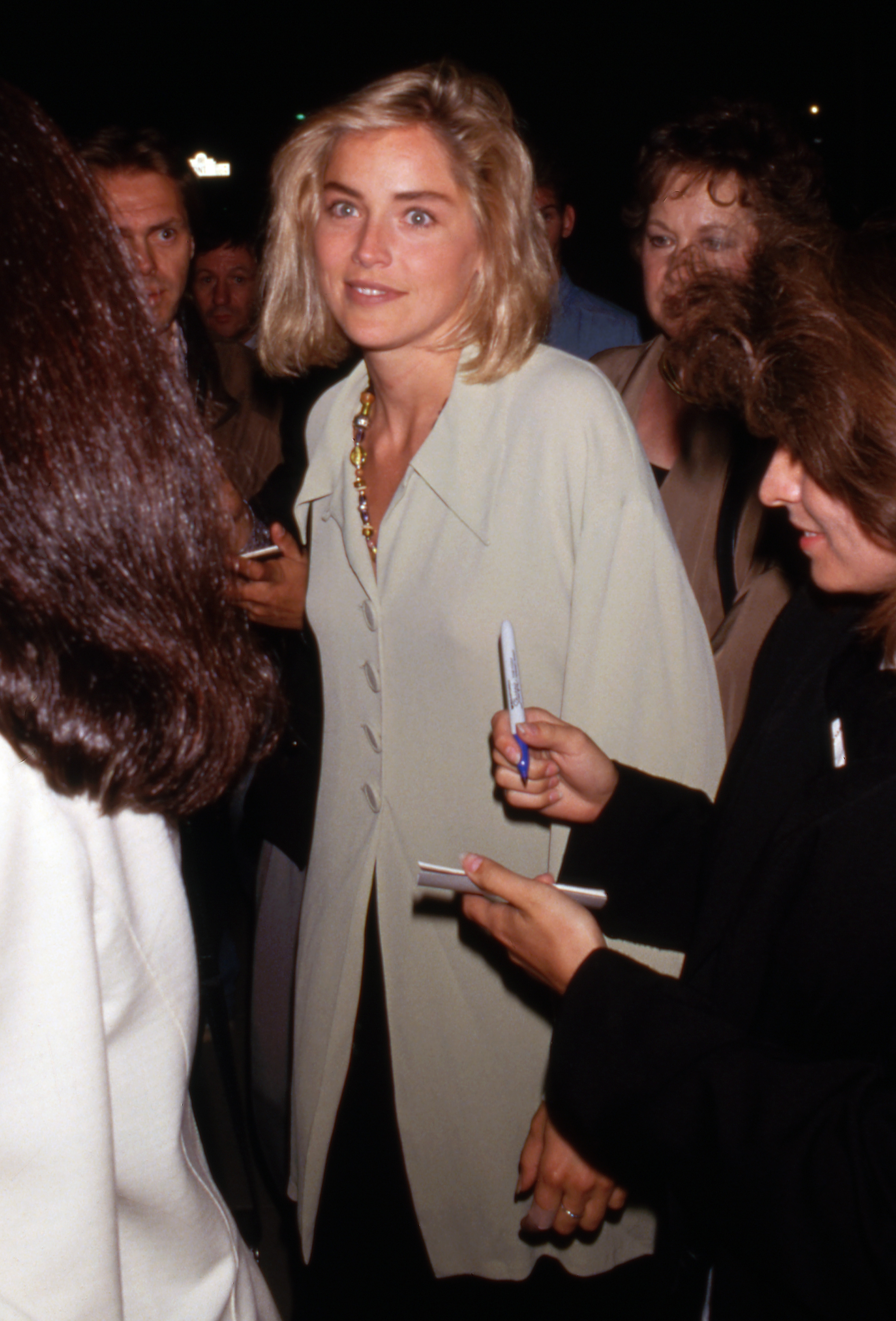 Sharon Stone looks on as she signs autographs in a crowd. Dressed in a pale oversized jacket with layered jewelry, she shows the fresh-faced style that marked her early public appearances.