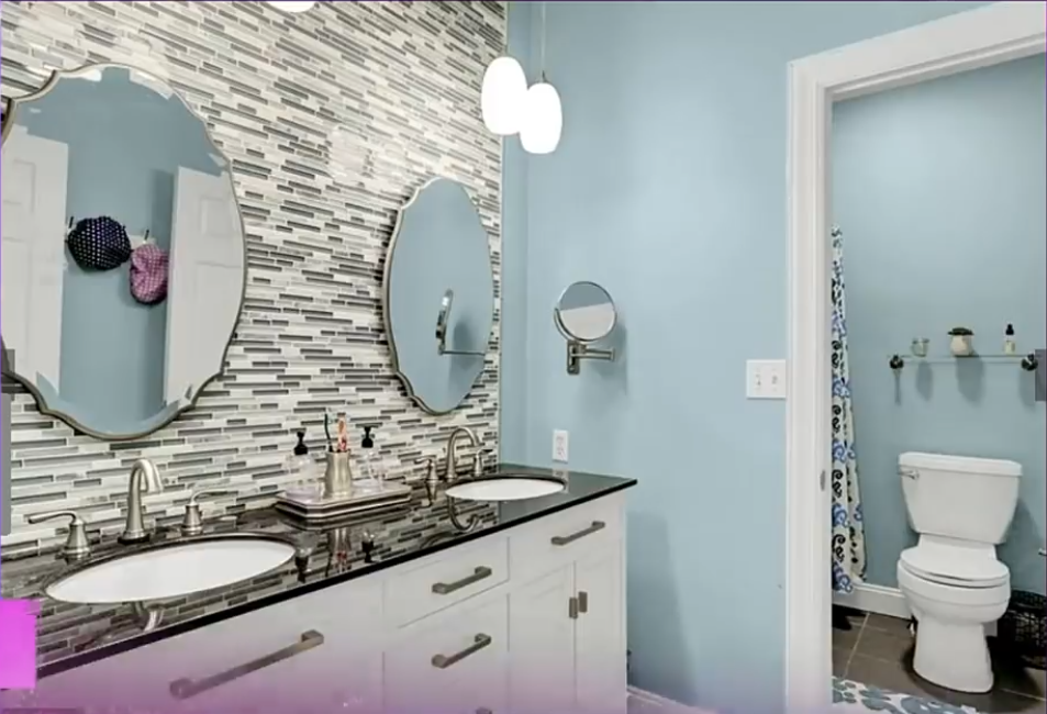 A double-sink vanity with white cabinetry and a dark countertop stretches beneath two decorative mirrors in this bathroom. Pale blue walls contrast with a mosaic-tile accent wall, while a separate toilet area is visible through the doorway.