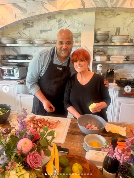 Wynonna smiles beside a man in an apron as they stand behind a counter covered with ingredients and flowers. The kitchen's open shelving and marble-patterned surfaces provide a detailed look at the cooking space. | Source: Instagram/wynonnajudd