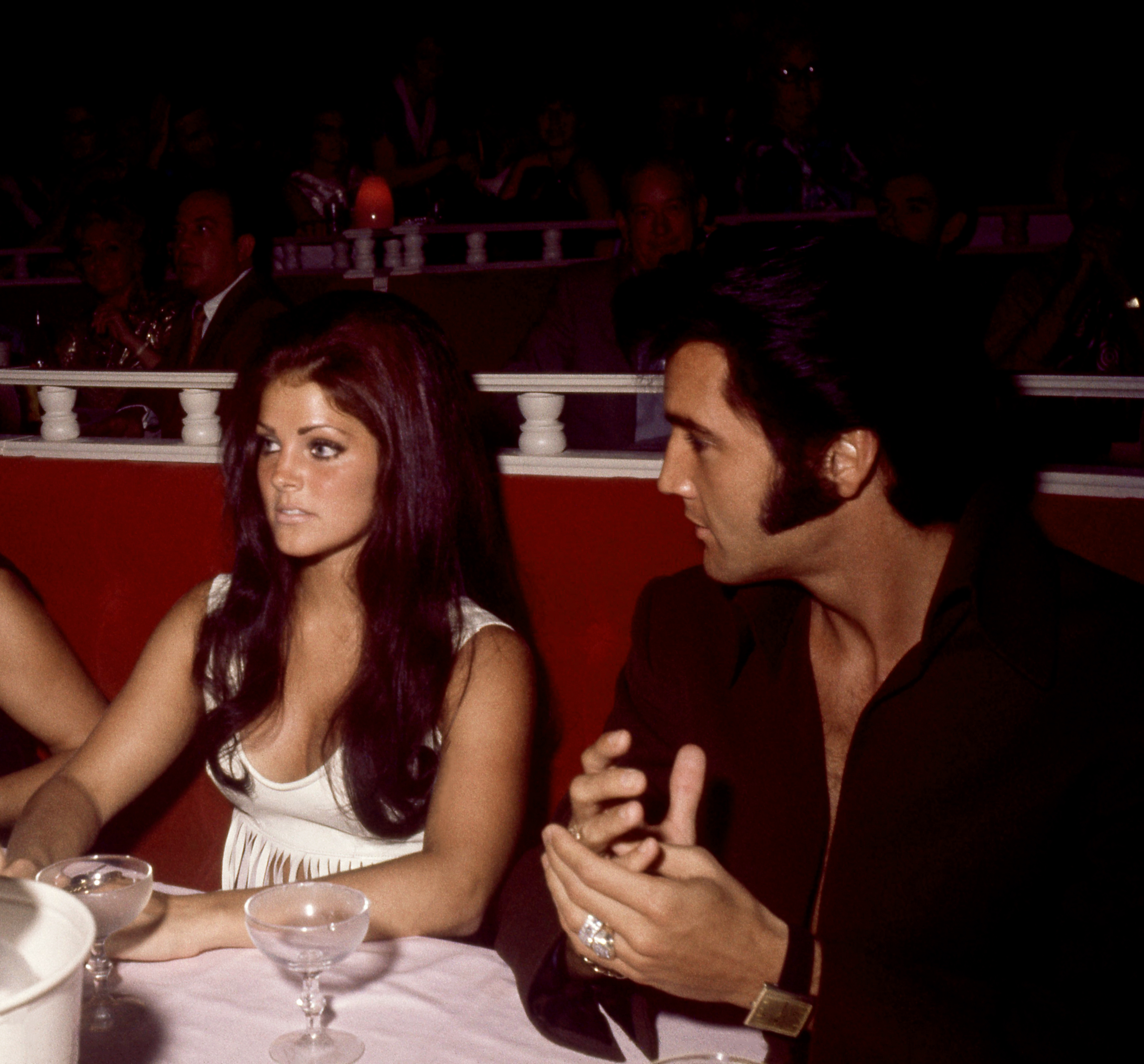 Priscilla Presley sits alongside husband Elvis Presley at a banquet table ahead of his opening performance at the International Hotel in Las Vegas, Nevada, in August 1969, wearing a sleeveless white scooped-neck top with her long dark hair styled in voluminous waves, beside Elvis dressed in a dark unbuttoned collared shirt. | Source: Getty Images