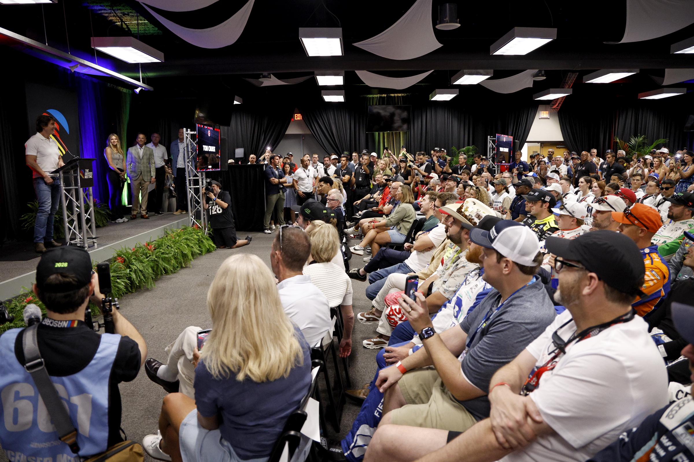Cruise addresses a packed room during the NASCAR gathering at Daytona International Speedway. The wide view shows him at the front as rows of drivers and other attendees listen from their seats. | Source: Getty Images