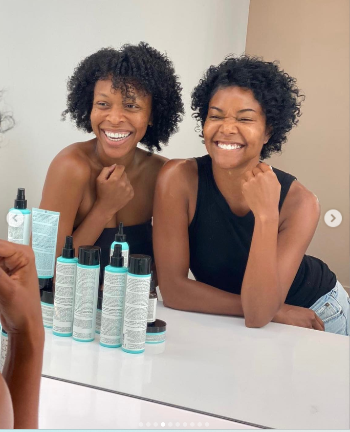 Gabrielle Union smiles widely beside another woman as they lean on a bathroom counter, their reflections visible in the mirror alongside neatly arranged haircare products. | Source: Instagram/gabunion