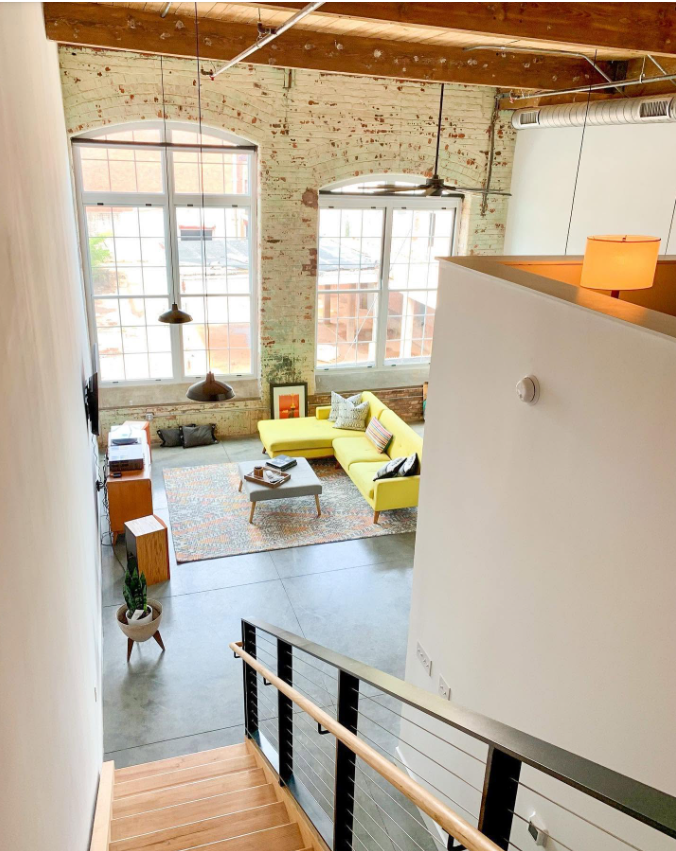 A view from the staircase opens onto the loft’s living area below, highlighting its multi-level design, soaring ceilings and expansive windows. | Source: Facebook/Judson Mills Loft