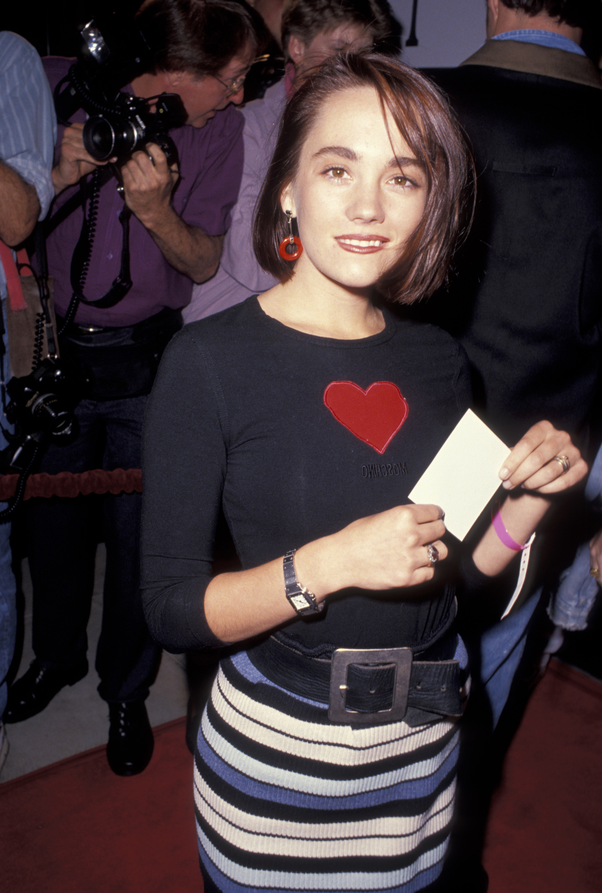 Natasha Gregson Wagner attends the Los Angeles premiere of "My Own Private Idaho" at the Academy Theater in Beverly Hills, circa October 1991. She wears a fitted black top with a red heart design and a striped skirt, accessorized with red earrings and a wide black belt.