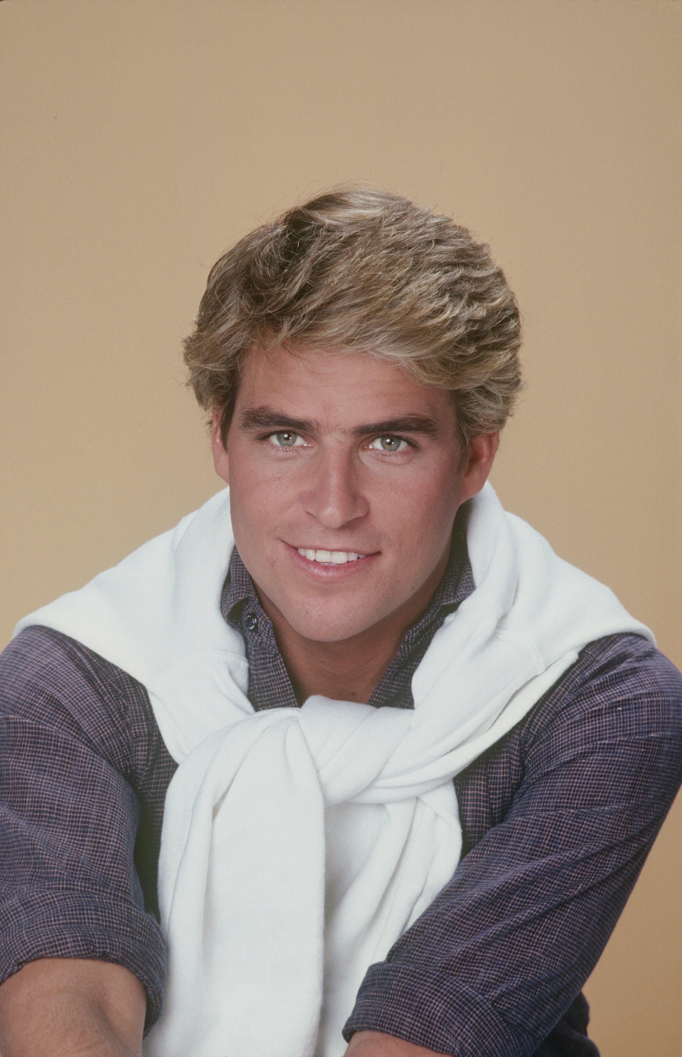 Ted McGinley flashed an all-American smile in a promotional portrait for the hit sitcom "Happy Days" on November 1, 1980. Channeling a quintessential 1980s preppy look, the young actor showed off feathered blonde hair while wearing a dark checkered button-down shirt paired with a white knit sweater casually draped and tied over his shoulders. | Source: Getty Images
