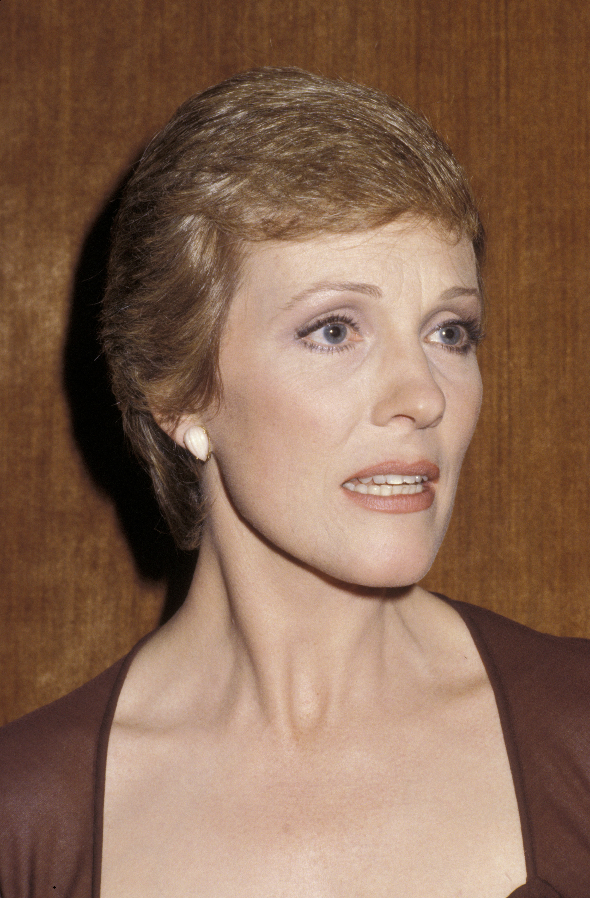 Julie Andrews attends the "Because We Care" benefit for Cambodia at the Dorothy Chandler Pavilion in Los Angeles on January 29, 1980. The actress exudes timeless poise in a close-up portrait, styled in a deep brown ensemble with delicate white shell-shaped stud earrings, soft evening makeup, and her signature cropped pixie cut.