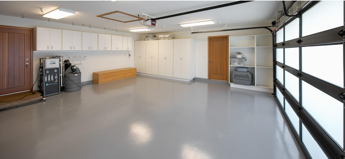 A spacious garage with storage cabinets. White built-in cabinets line the walls, while wooden doors and a wide translucent-paneled garage door add to the bright interior. Equipment and storage units occupy one side, leaving a generous amount of open floor space. | Source: Redfin