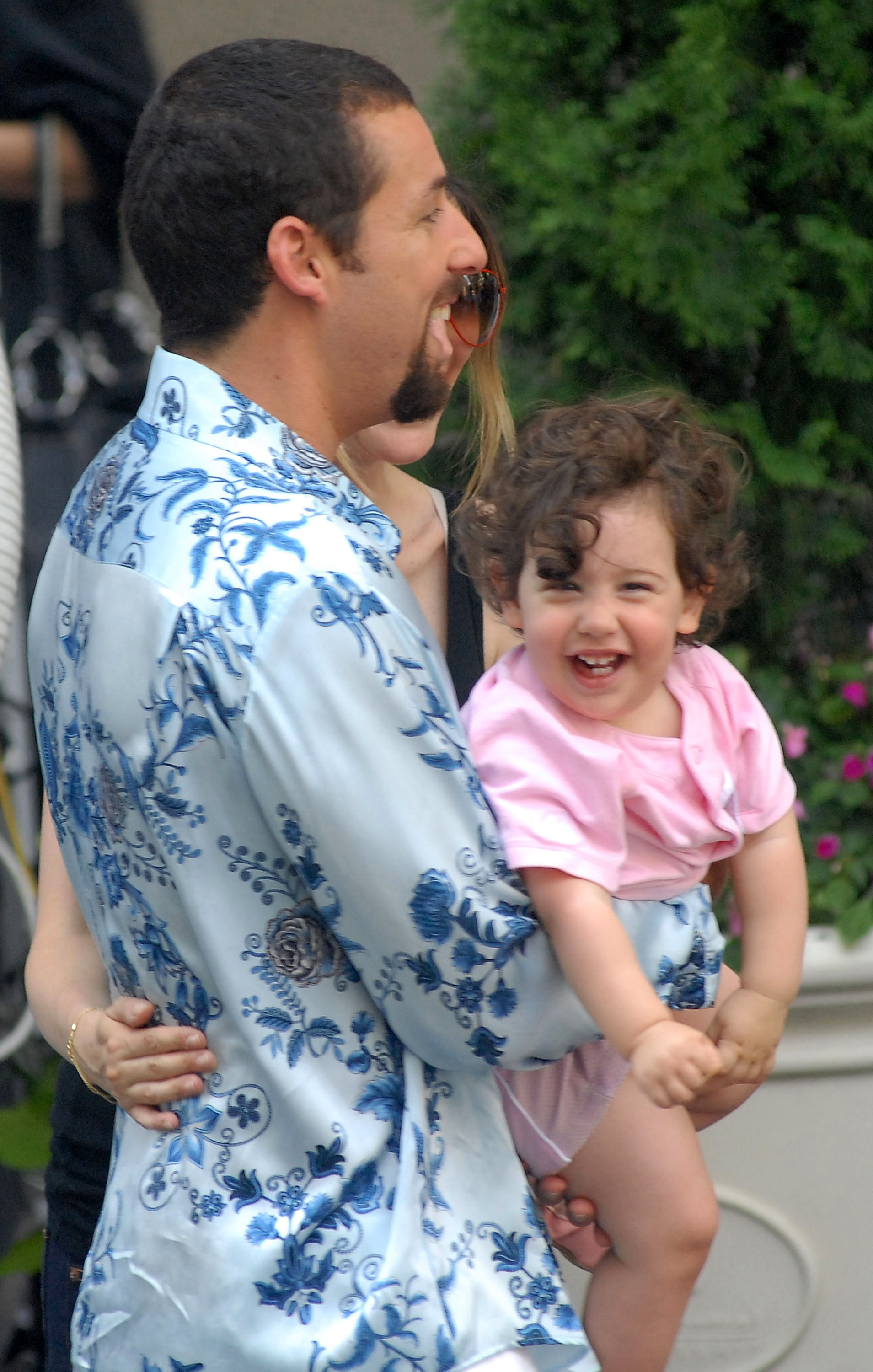Adam Sandler holds his one-year-old daughter Sadie in his arms, the toddler beaming with joy as she looks toward the camera. Dressed casually on set, the candid moment captures a softer side of the actor — playful, relaxed, and fully immersed in fatherhood.