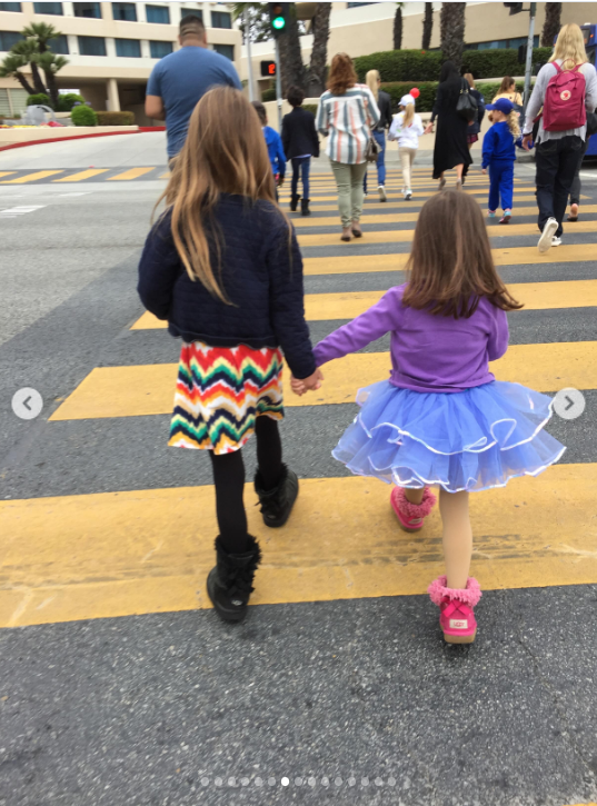 them: their hands clasped together as they walk. Mabel and Fiona are pictured from behind as they hold hands while crossing a yellow-striped pedestrian crosswalk. Mabel wears a dark jacket, colorful zigzag-patterned skirt and black boots, while Fiona wears a purple top, bright blue tutu-style skirt and pink shoes, capturing a sweet moment of the sisters walking side by side. | Source: Instagram/mabellowe_