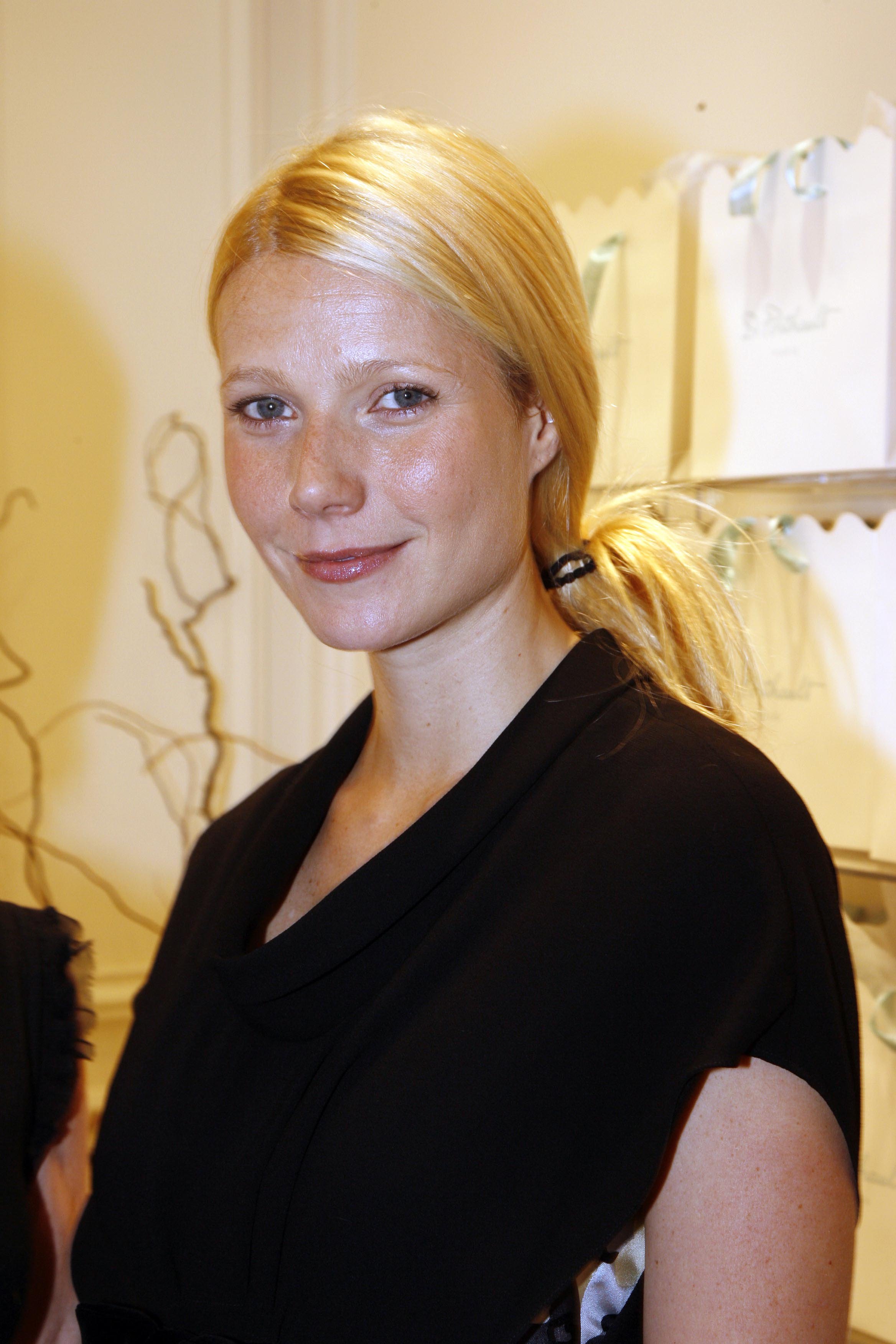 Gwyneth Paltrow attends a Paris Fashion Week party in France on October 5, 2006. She wears a structured black dress with a modern draped neckline, while her blonde hair is gathered into a low ponytail. A subtle sheen across her complexion and a hint of gloss give her look a polished yet understated finish.