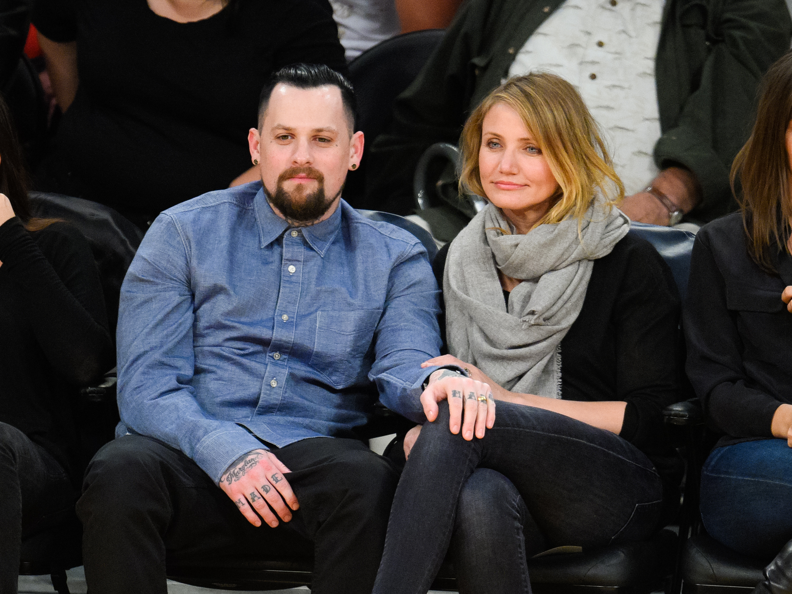 Benji Madden and Cameron Diaz sit courtside at a basketball game at Staples Center, dressed casually, with Diaz in a dark outfit and gray scarf, leaning in close beside Madden as they watch the game.