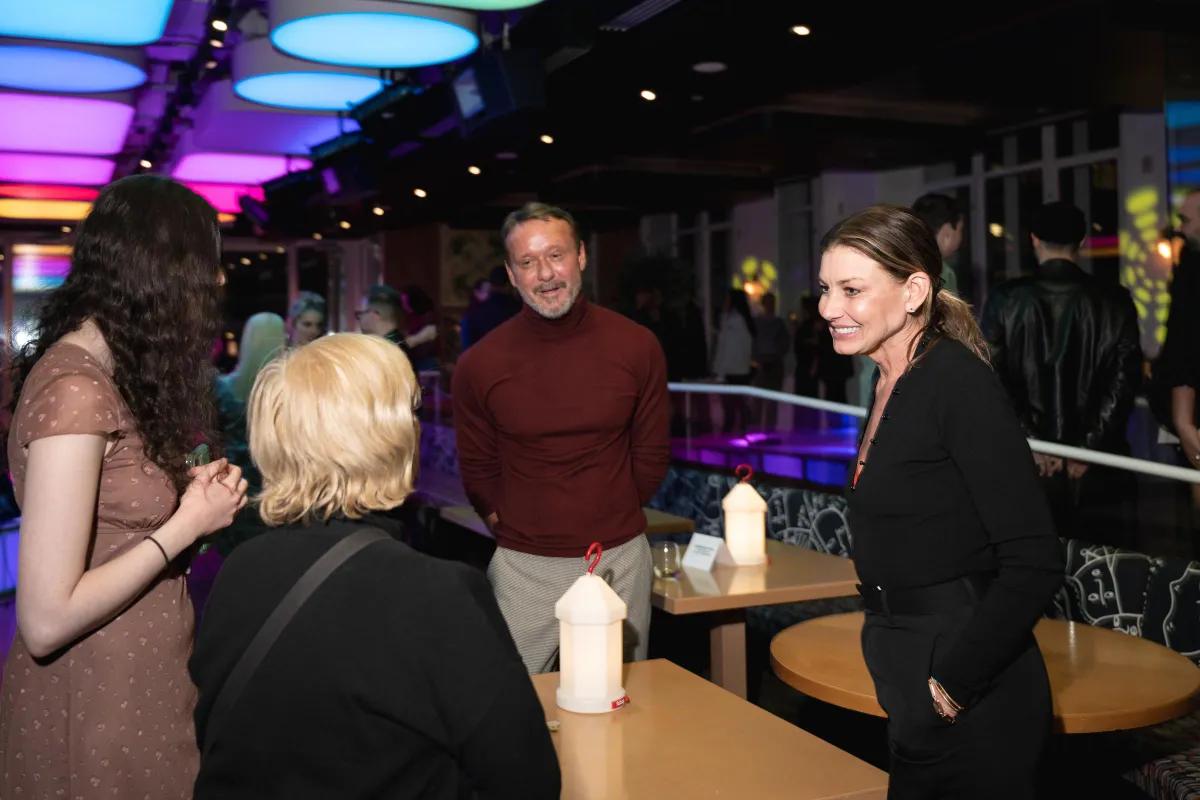 At the opening celebration for "Babe" at The Green Fig, Faith Hill and Tim McGraw are caught mid-conversation with guests beneath moody lighting. The candid moment feels unguarded, natural, and quietly intimate.
