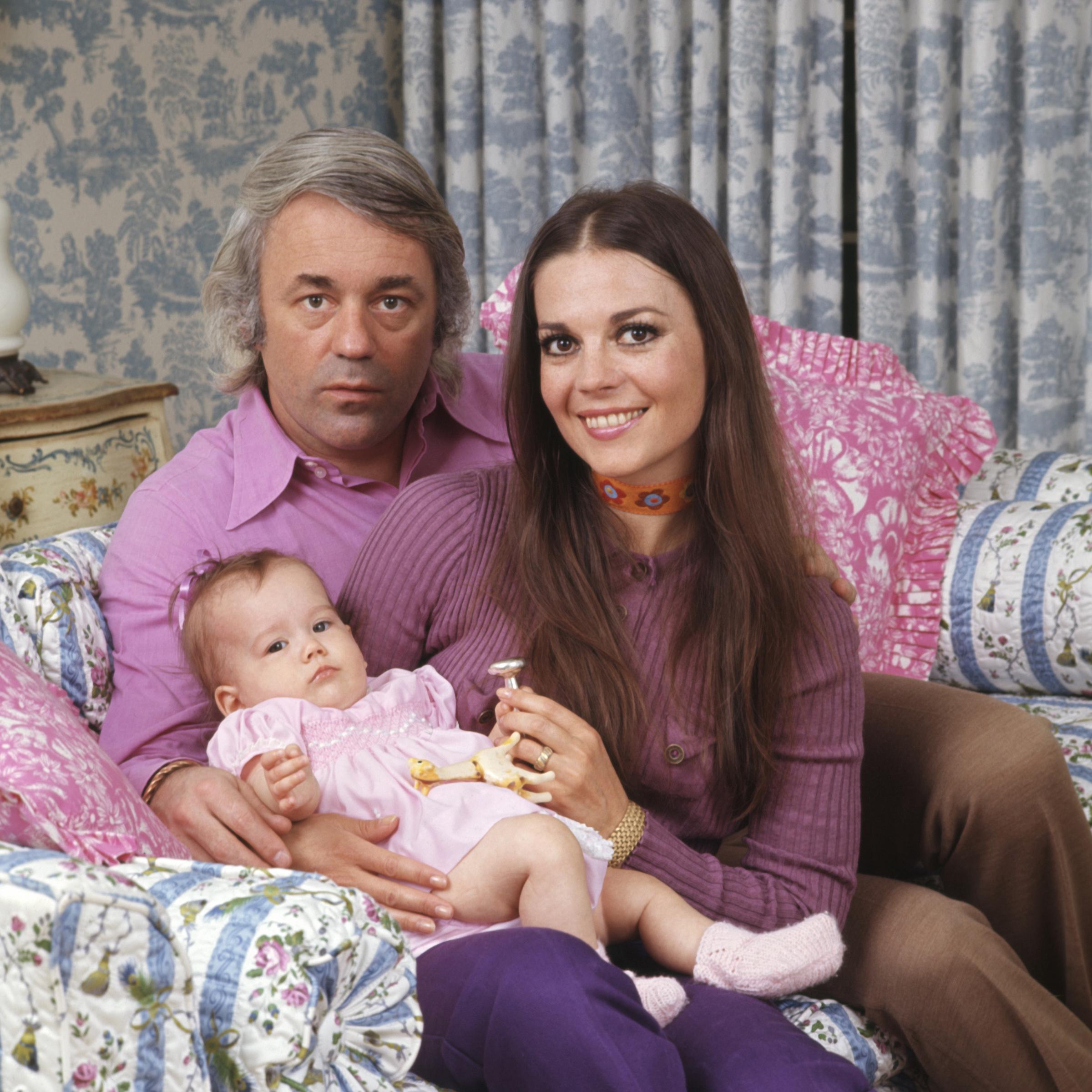 Natalie Wood sits beside her husband Richard Gregson, holding their baby daughter Natasha Gregson Wagner in her arms. Natalie wears a purple ribbed top and violet pants, accessorized with a colorful floral choker. Richard is dressed in a light pink shirt and beige trousers. Baby Natasha wears a pale pink dress with puffed sleeves and a tiny bow in her hair, resting calmly with a toy in her hand.