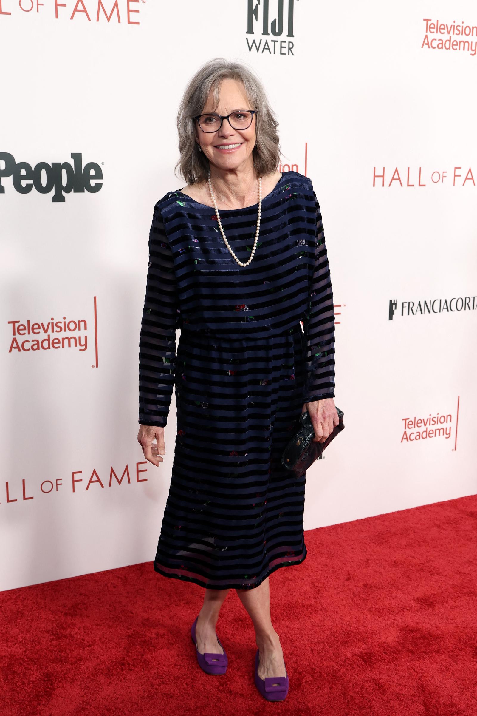 A full shot captures Sally Field glowing in a dark blue dress with black stripes, accessorized with her signature pearl necklace. She strikes a graceful pose with her legs crossed and a beautiful smile lighting up her face.
