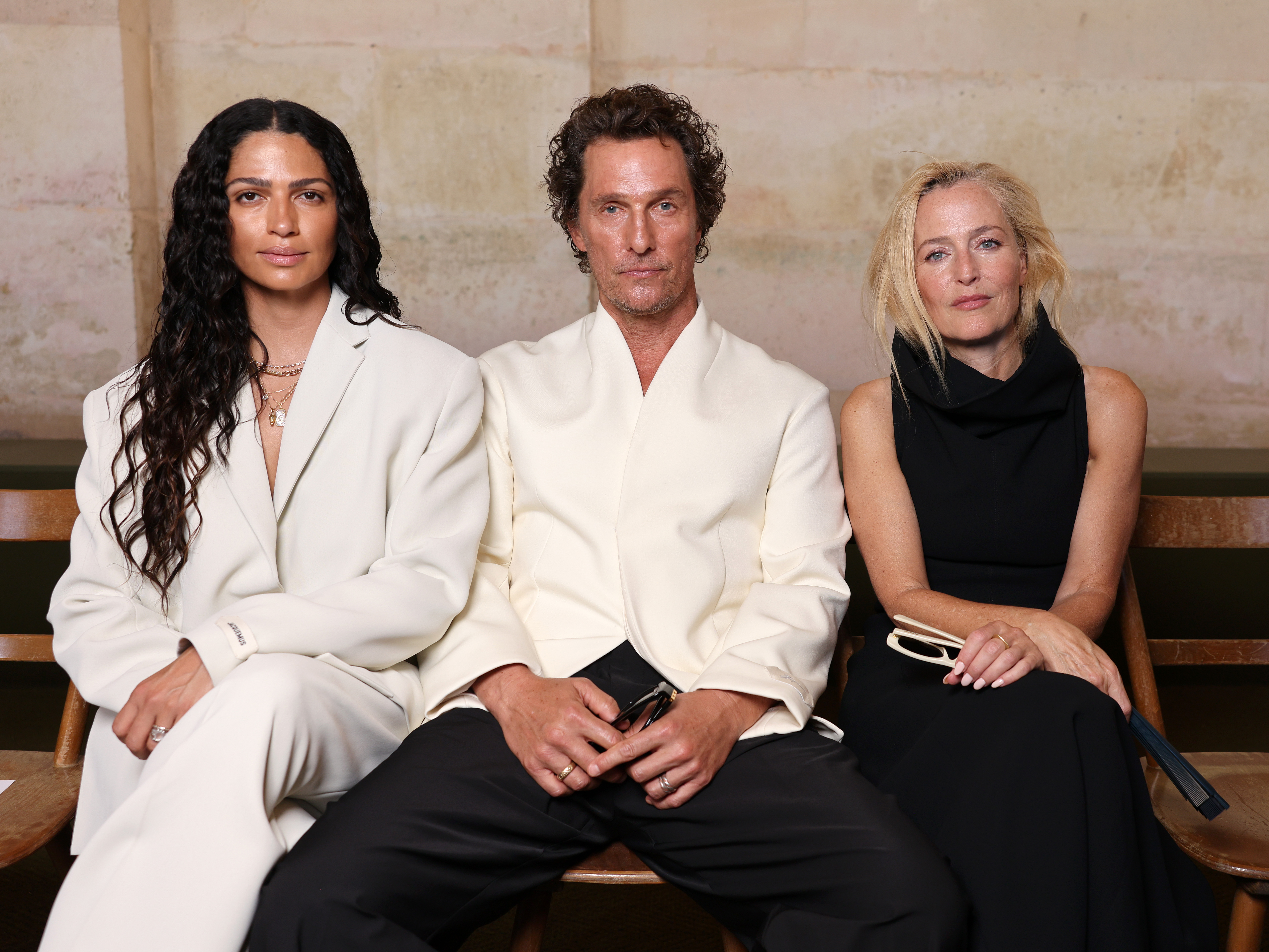 Alves, McConaughey, and Gillian Anderson are captured seated together during the Jacquemus fashion show. McConaughey sits sandwiched between the two women, all three facing the camera with composed, calm expressions.