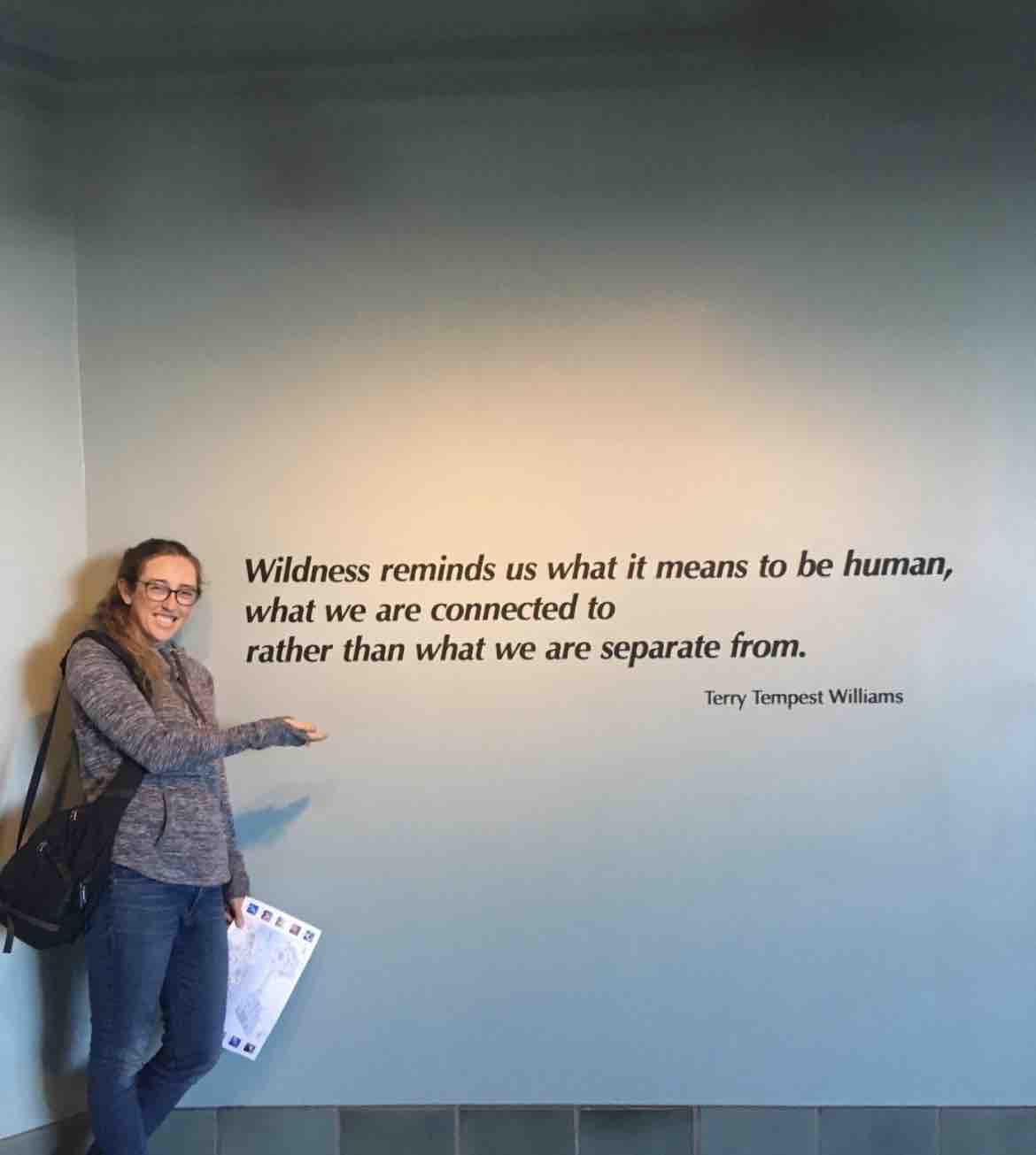 Dr. Julia Pederson gestures toward a museum wall inscribed with a Terry Tempest Williams quote on human connection to nature, holding a visitor guide and carrying a shoulder bag. Outside of her demanding medical career, she found joy and restoration in visiting local nature preserves, reading, and learning to crochet, embracing life’s quieter passions alongside her devoted work with young families. | Source: GoFundMe