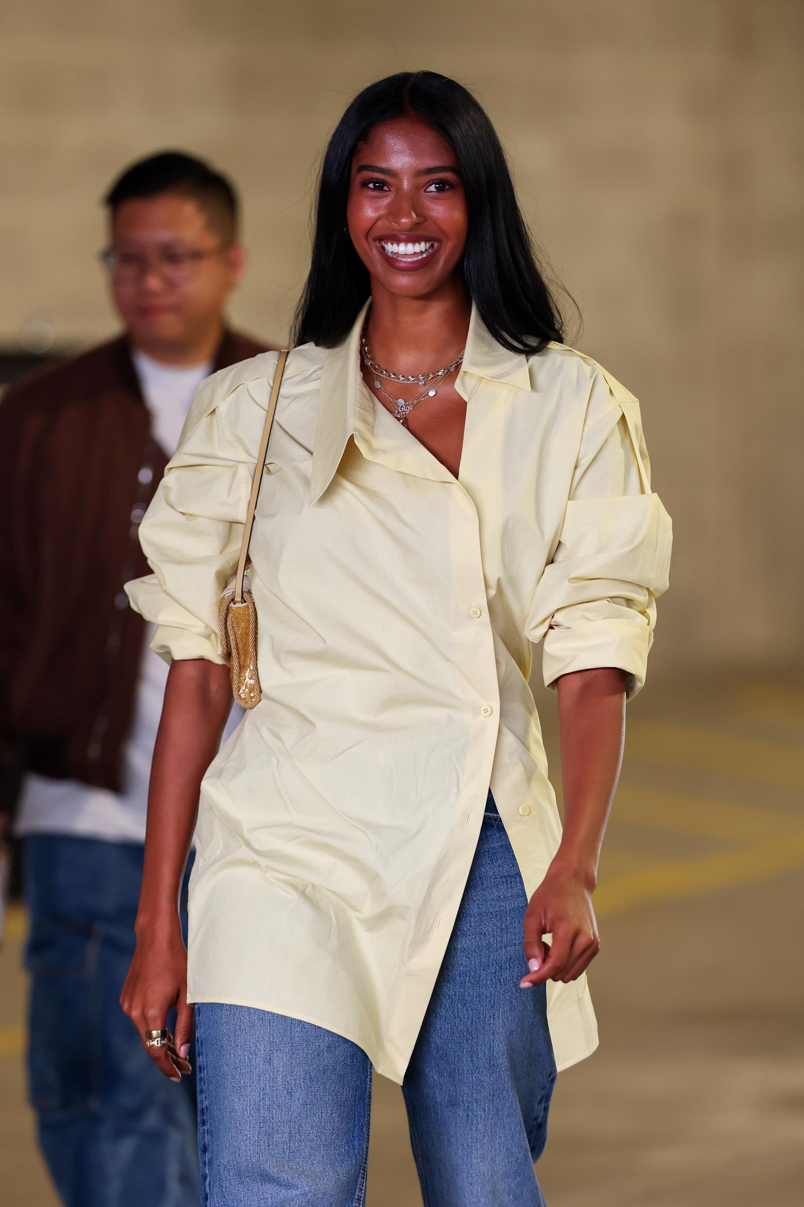 Natalia Bryant arrives on the blue carpet during Day Ten of the 2026 US Open, wearing her pale-yellow jacket with wide-leg jeans and a gold-toned shoulder bag as she smiles toward photographers.