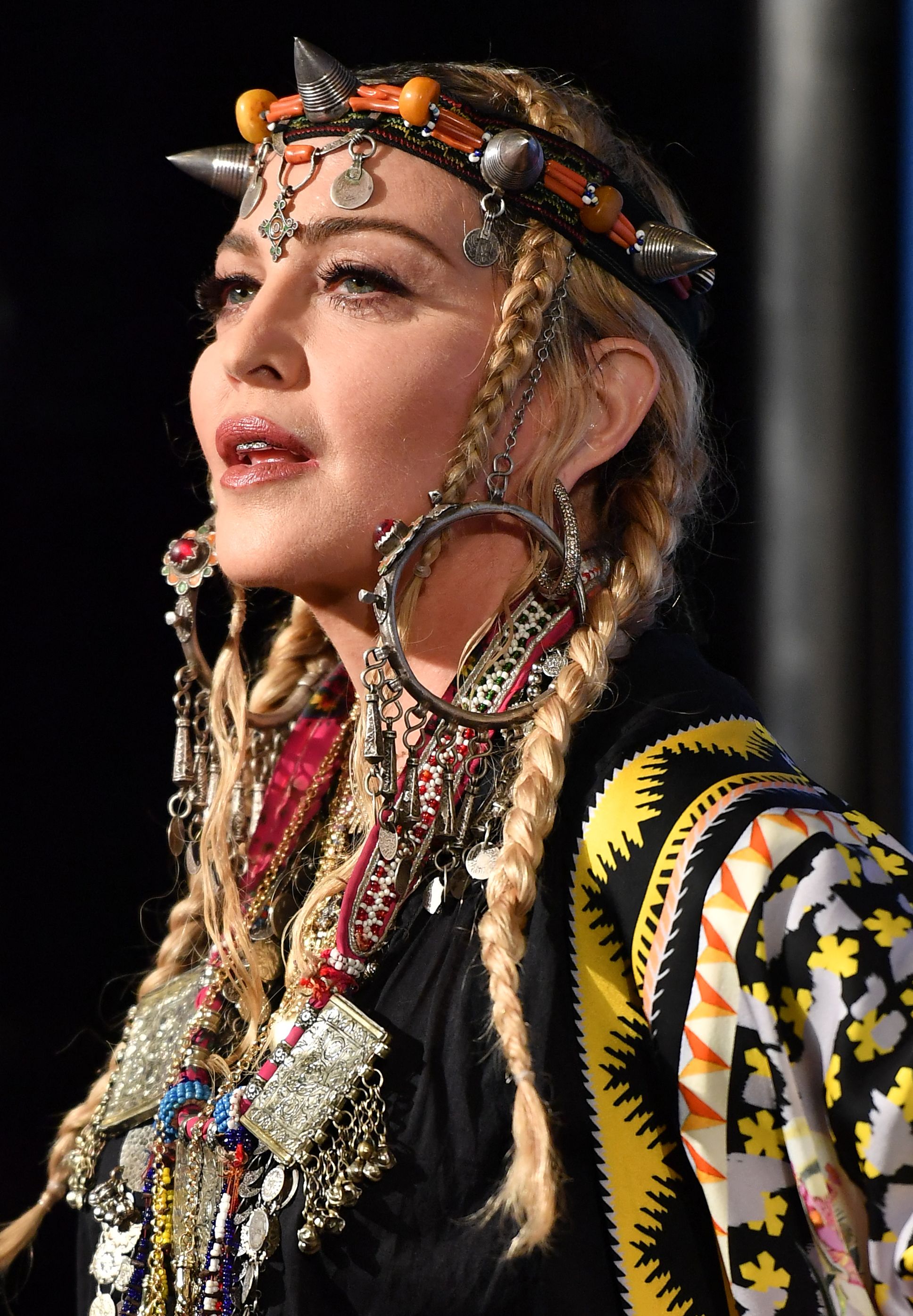 Madonna is photographed from the side wearing long blond braids, a decorative headpiece, oversized earrings and layers of intricate jewelry. | Source: Chat GPT