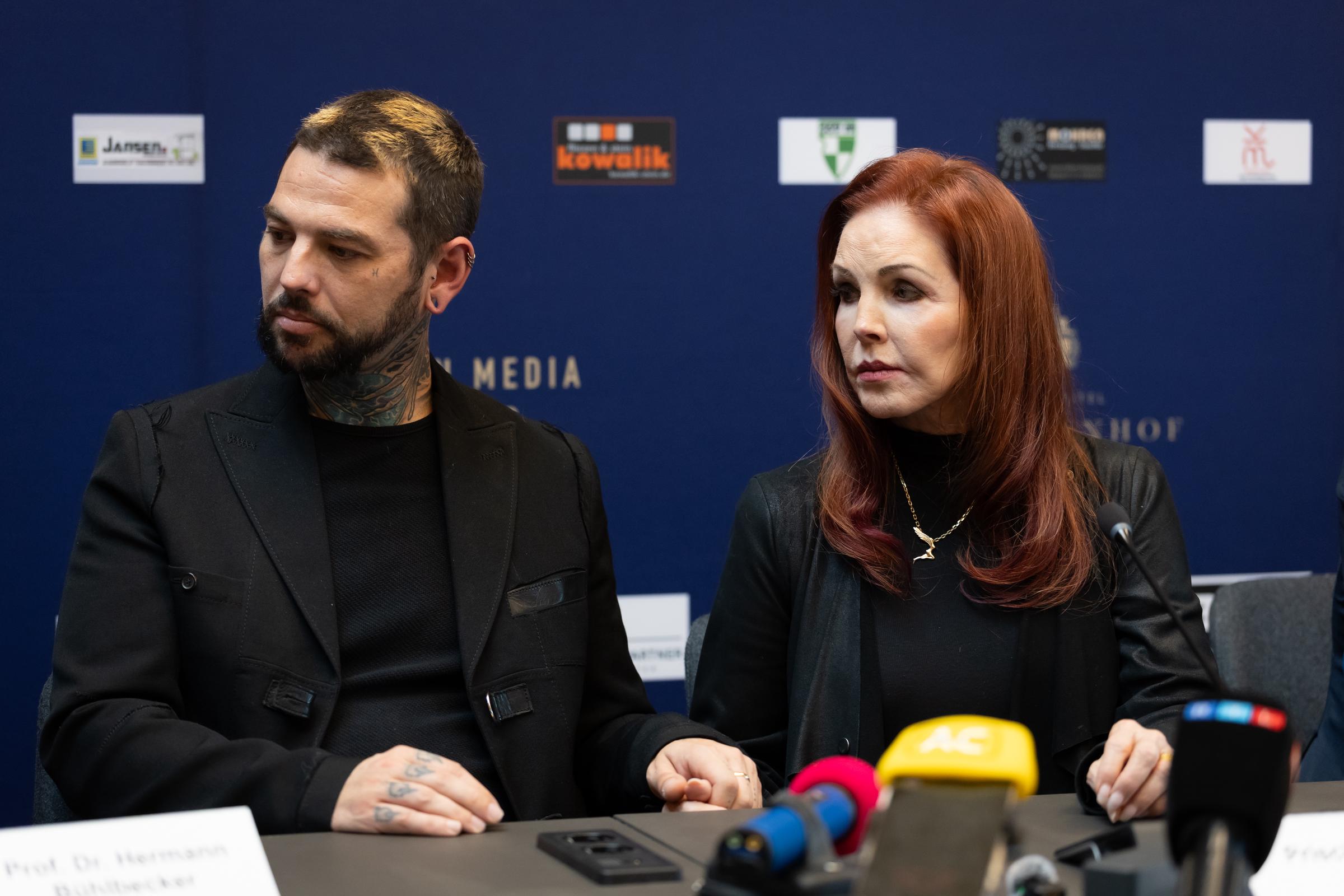 Priscilla Presley sits alongside her son, Navarone Garibaldi Garcia, during a press event in Baesweiler, Germany, on October 20, 2026, both dressed in coordinated black attire as they listen intently behind microphones at a panel table, with Presley wearing a gold necklace over a black top and styling her layered auburn hair in soft curves around her shoulders.