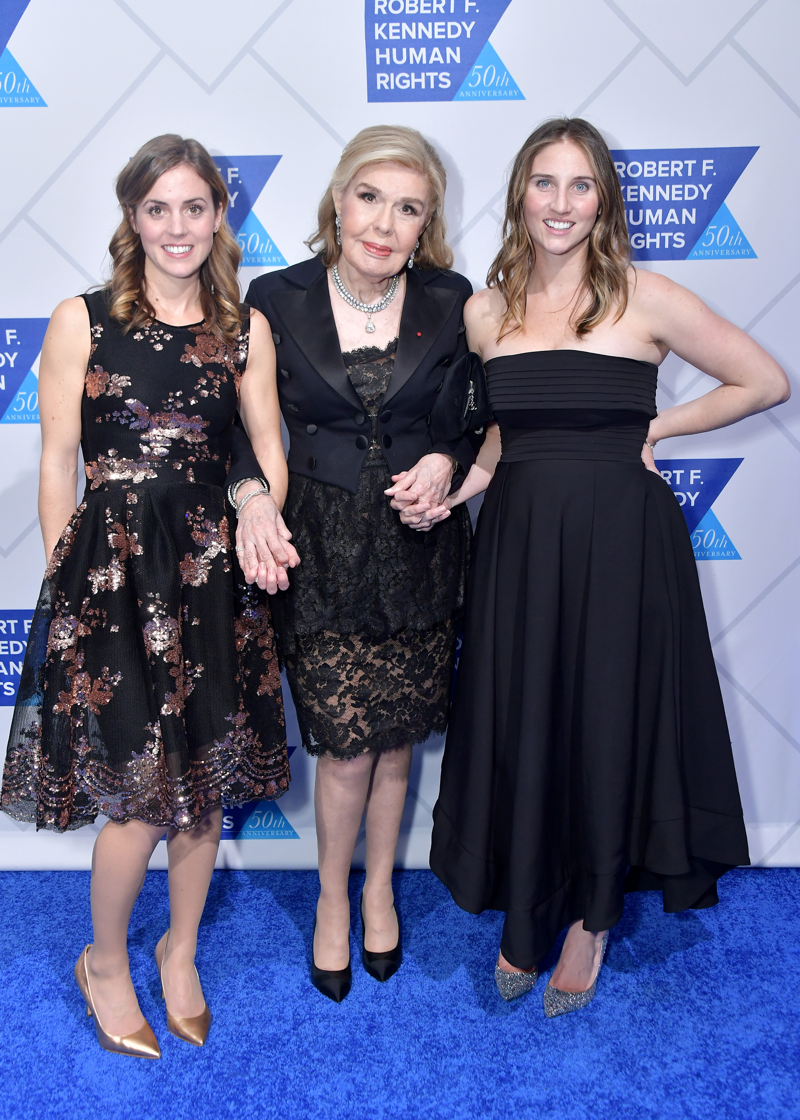 Kyle Kennedy Kerr, Marianna Vardinoyannis, and Rory Kennedy Camillo link hands on the blue carpet at the 2019 Robert F. Kennedy Human Rights Ripple of Hope Awards on December 12, 2018, in New York City. Kyle wears a sequined knee-length black dress with metallic pumps, standing alongside Marianna in an elegant black lace ensemble with a cropped jacket and pearl necklace, while Rory poses on the right in a strapless black pleated gown. | Source: Getty Images