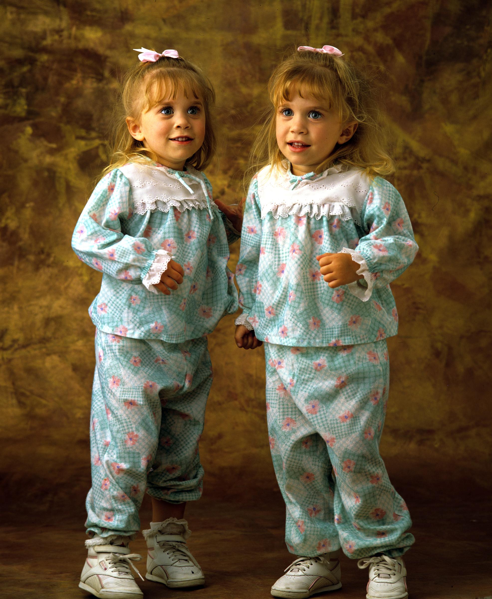Four-year-old twins traded toddler soft-focus for alert, expressive little-kid faces framed by blunt bangs on September 21, 1990, channeling early 1990s comfort in coordinated turquoise and soft pink loungewear sets with eyelet lace bibs, ruffled cuffs, and white high-top sneakers.